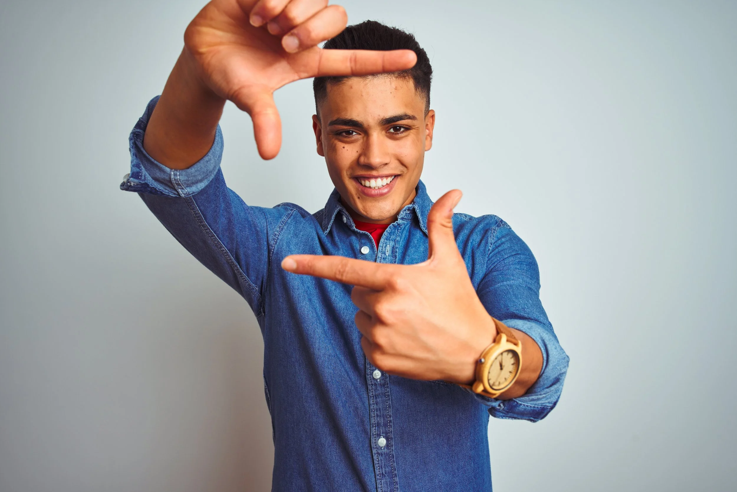 Young man smiling, making a rectangular frame with his hands, wearing a denim shirt and a watch on his left wrist.