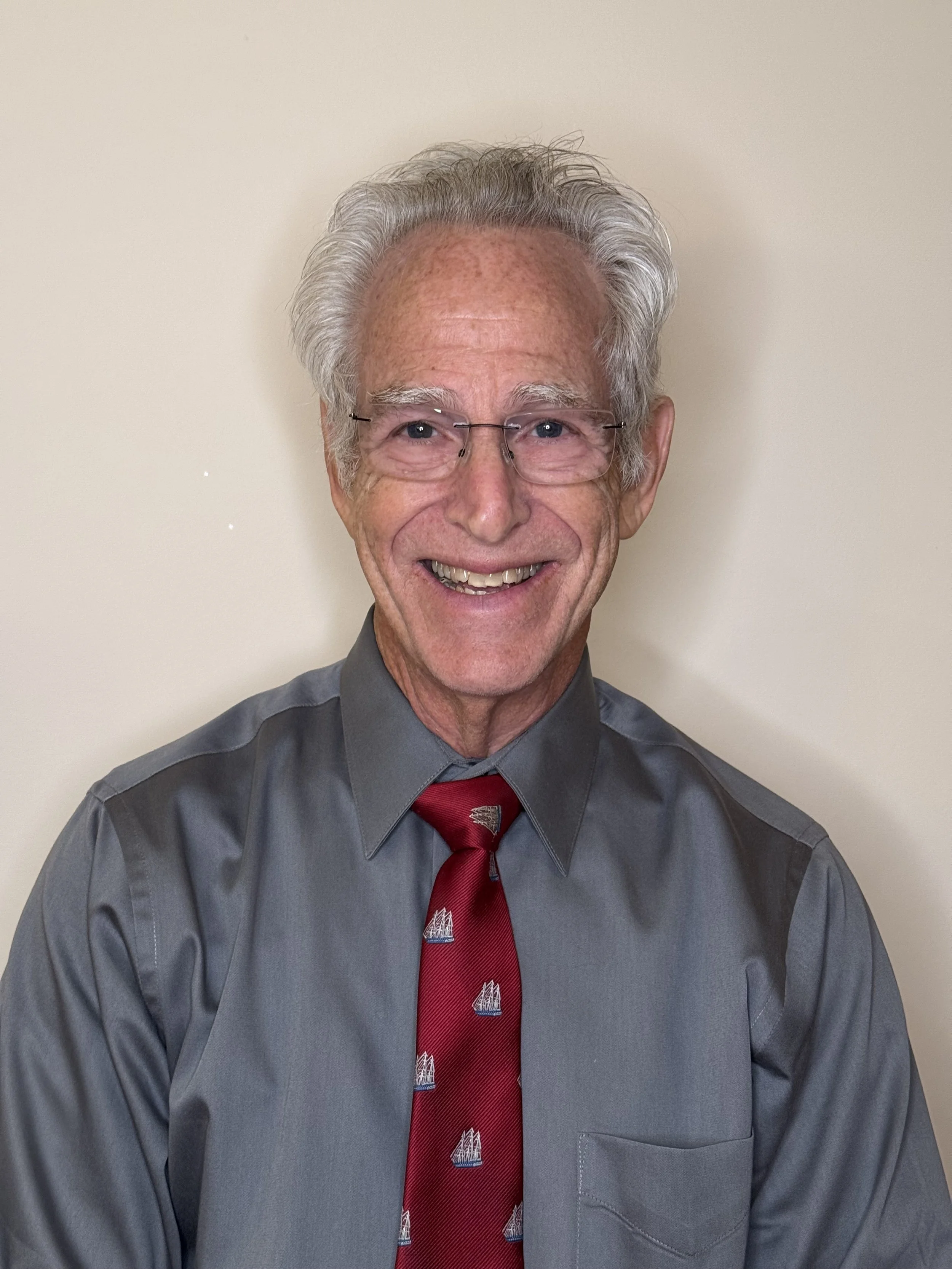 A smiling man with white hair and glasses, wearing a gray dress shirt and a red tie standing against a plain beige wall.