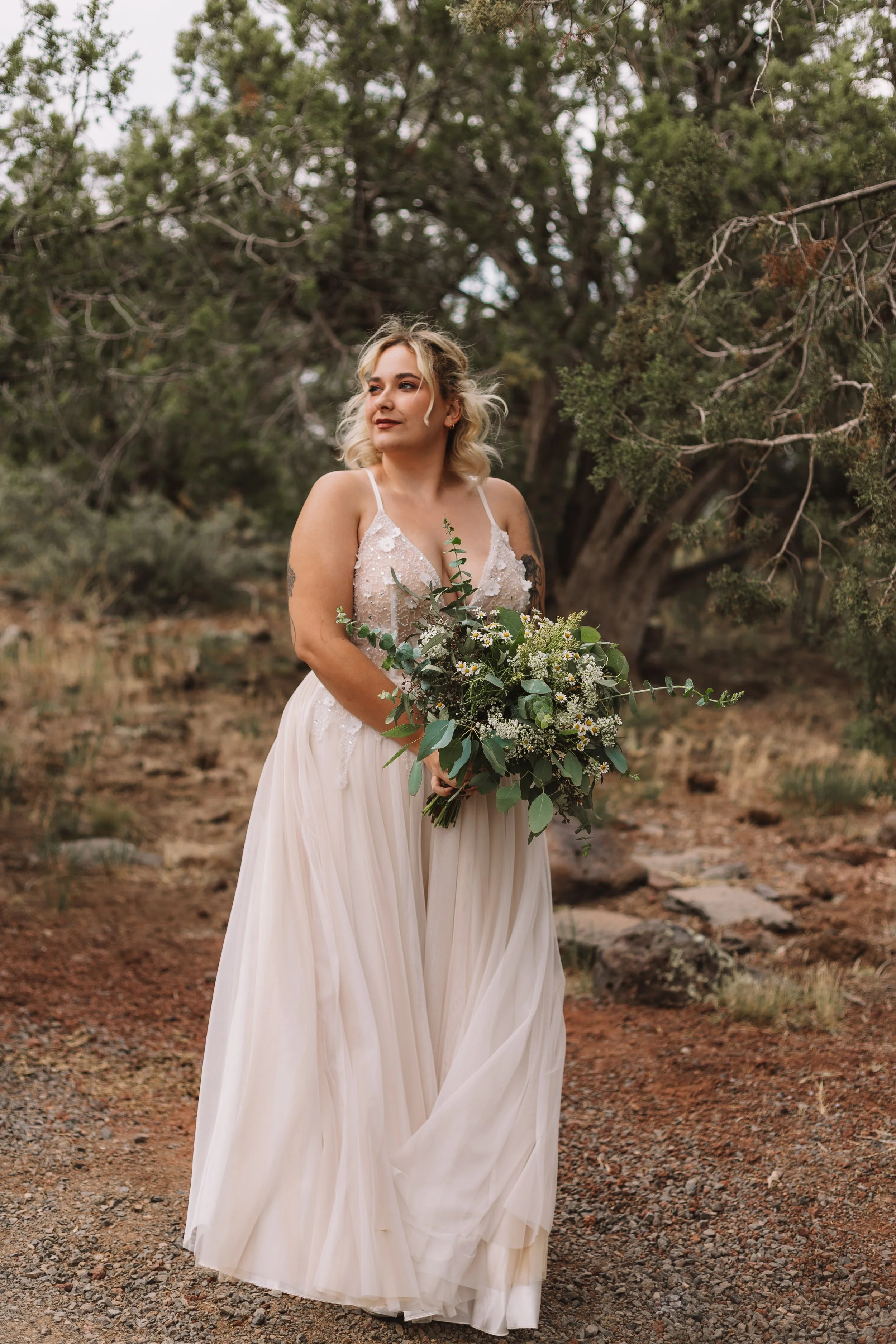 Bride in white dress holding a bouquet in a natural outdoor setting with trees.