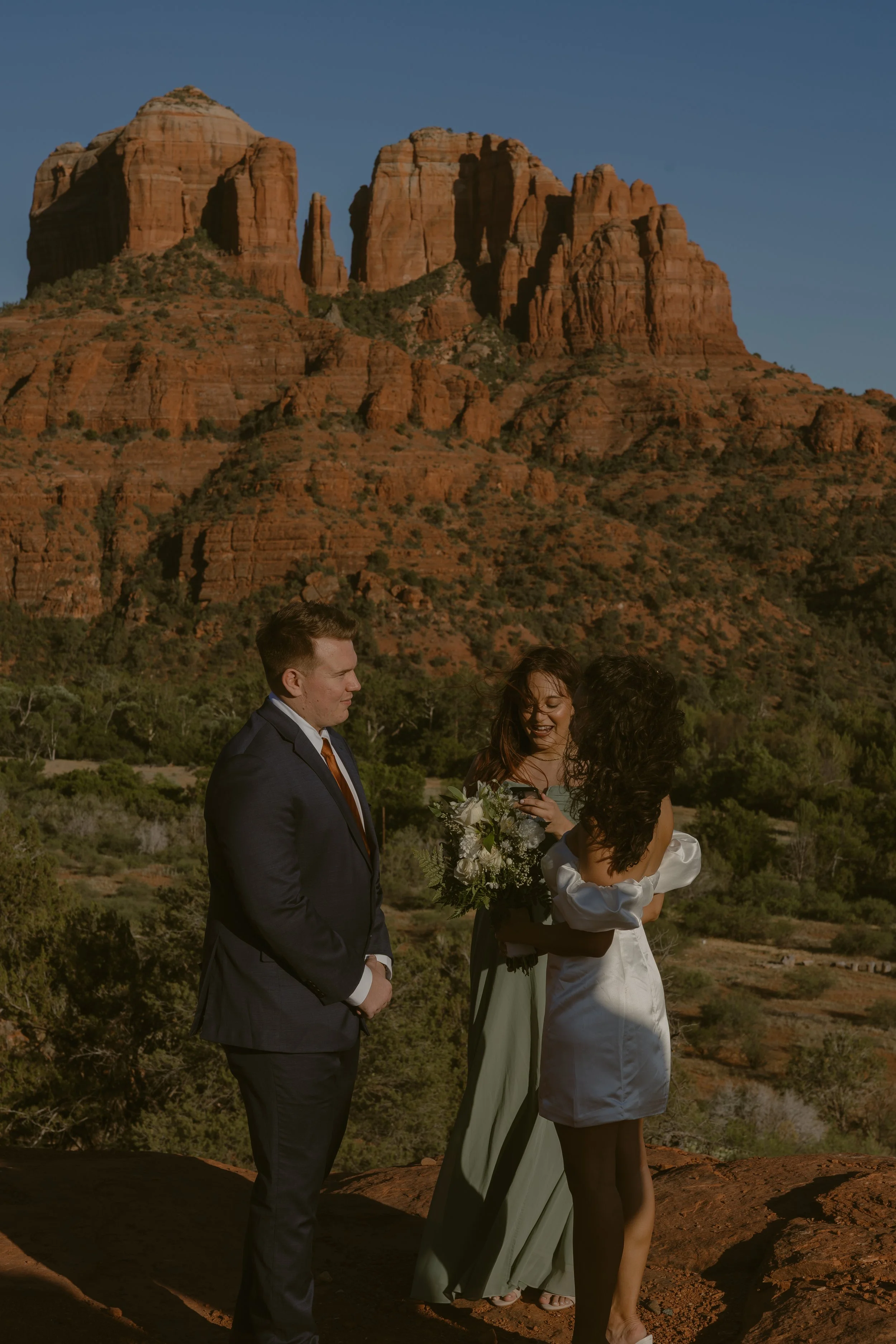 A wedding ceremony taking place outdoors with red rock mountains in the background, featuring a bride, groom, and officiant.