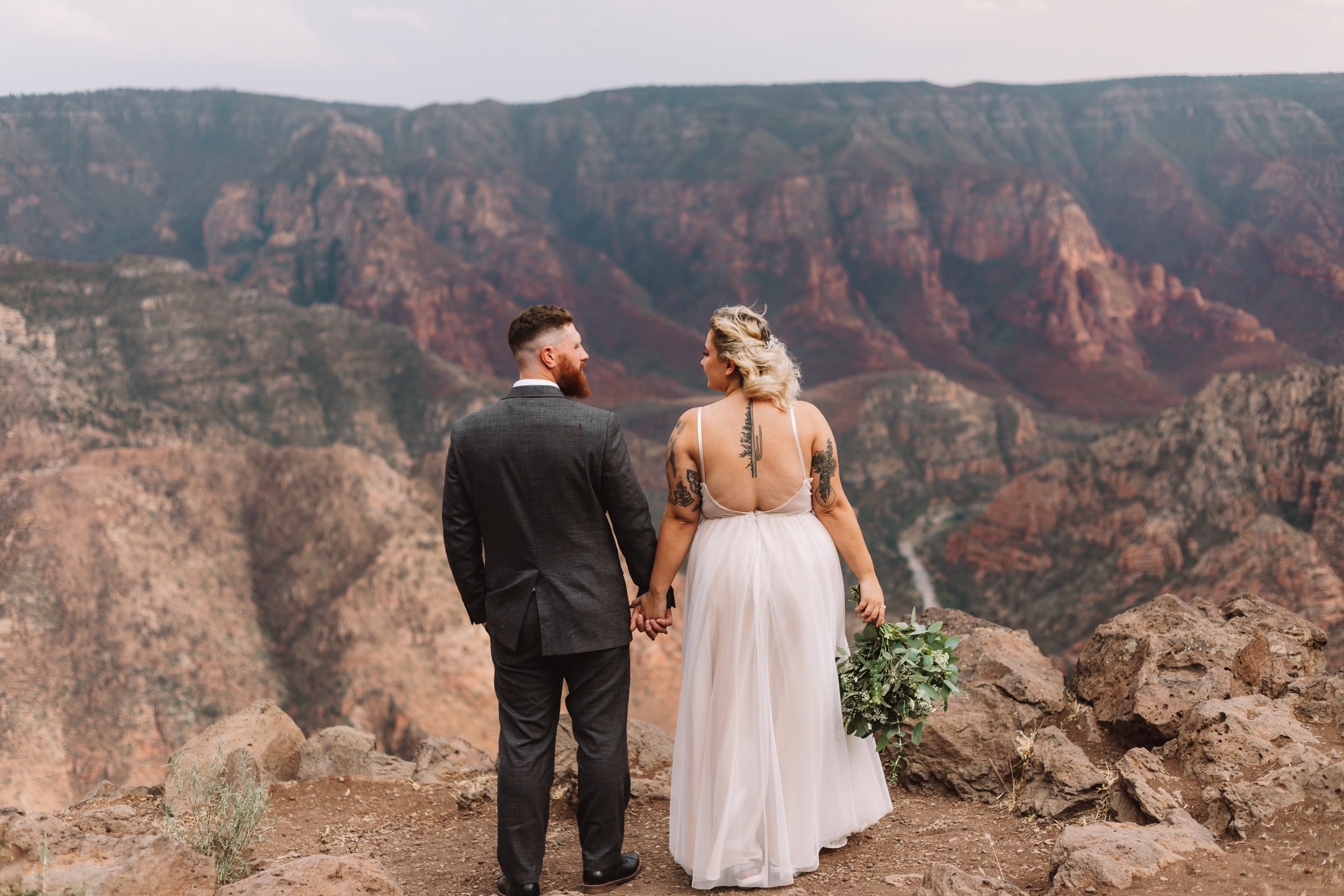 Bride and groom holding hands, standing on rocky cliff with canyon view, wedding attire.