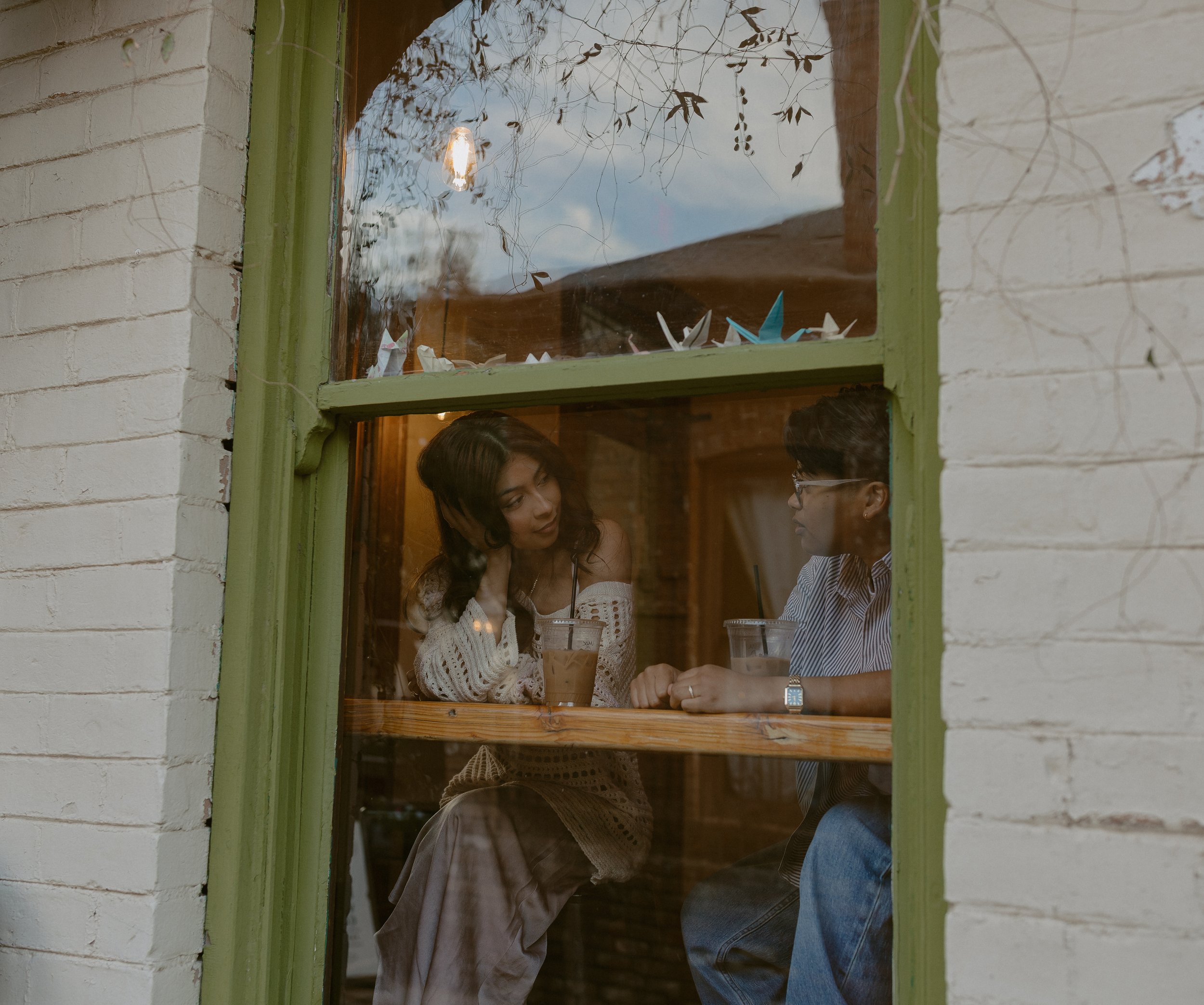 Two people sitting at a table inside a cafe seen through a window. The woman on the left has long dark hair, is wearing a white knitted sweater and a skirt, and the man on the right has short dark hair, glasses, and is wearing a striped shirt. Both h