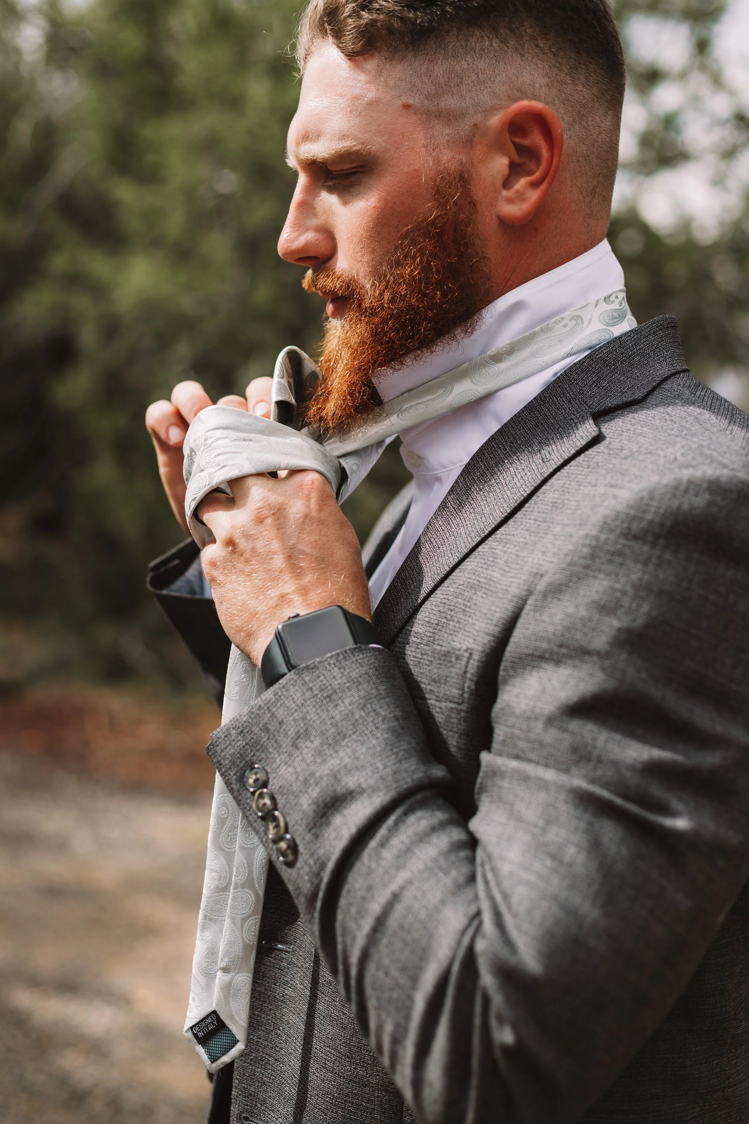 Man in a gray suit tying a paisley-patterned tie outdoors.