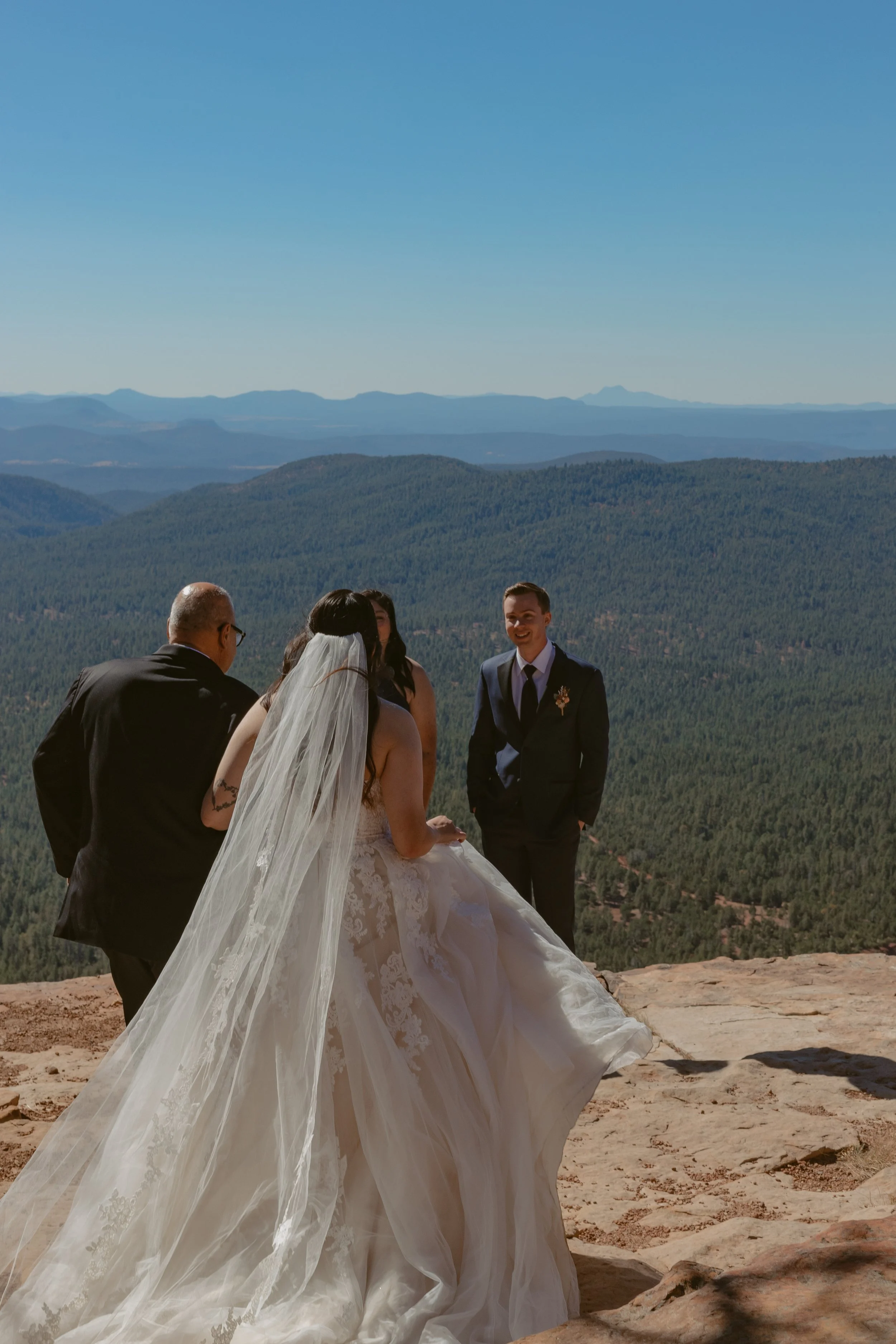 Two people sitting at an outdoor glass table, viewed from a window, with drinks and a notebook, in a brick-paved patio surrounded by greenery and brick walls.