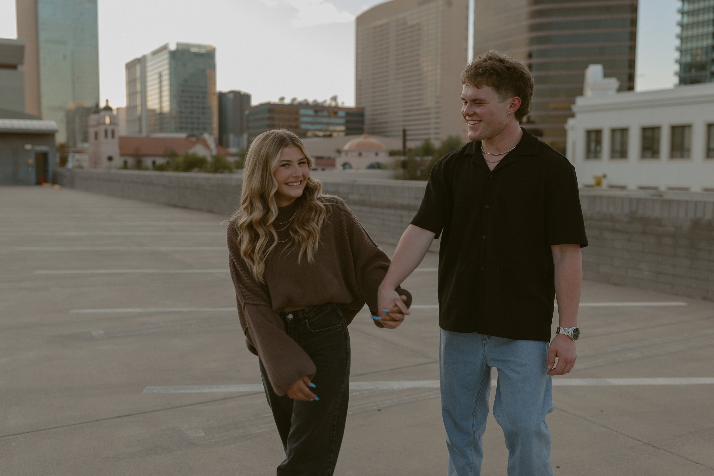 A young couple holding hands and smiling on a rooftop with city buildings in the background.