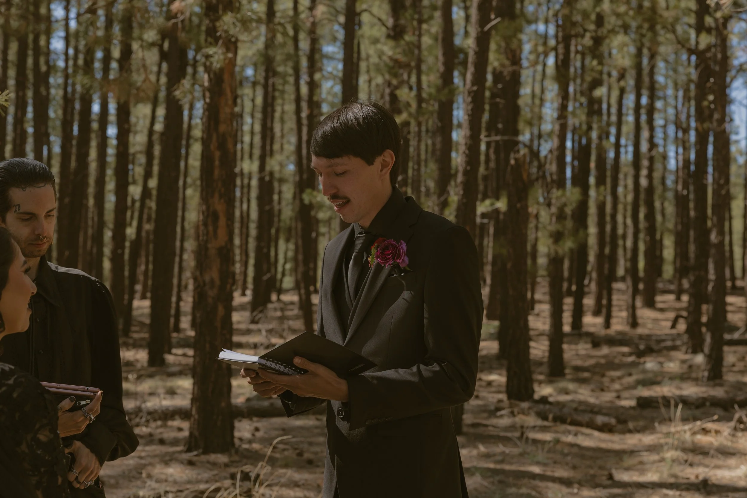 A groom in a black suit with a purple boutonniere stands in a forest during a wedding ceremony, holding a booklet, with guests around him.