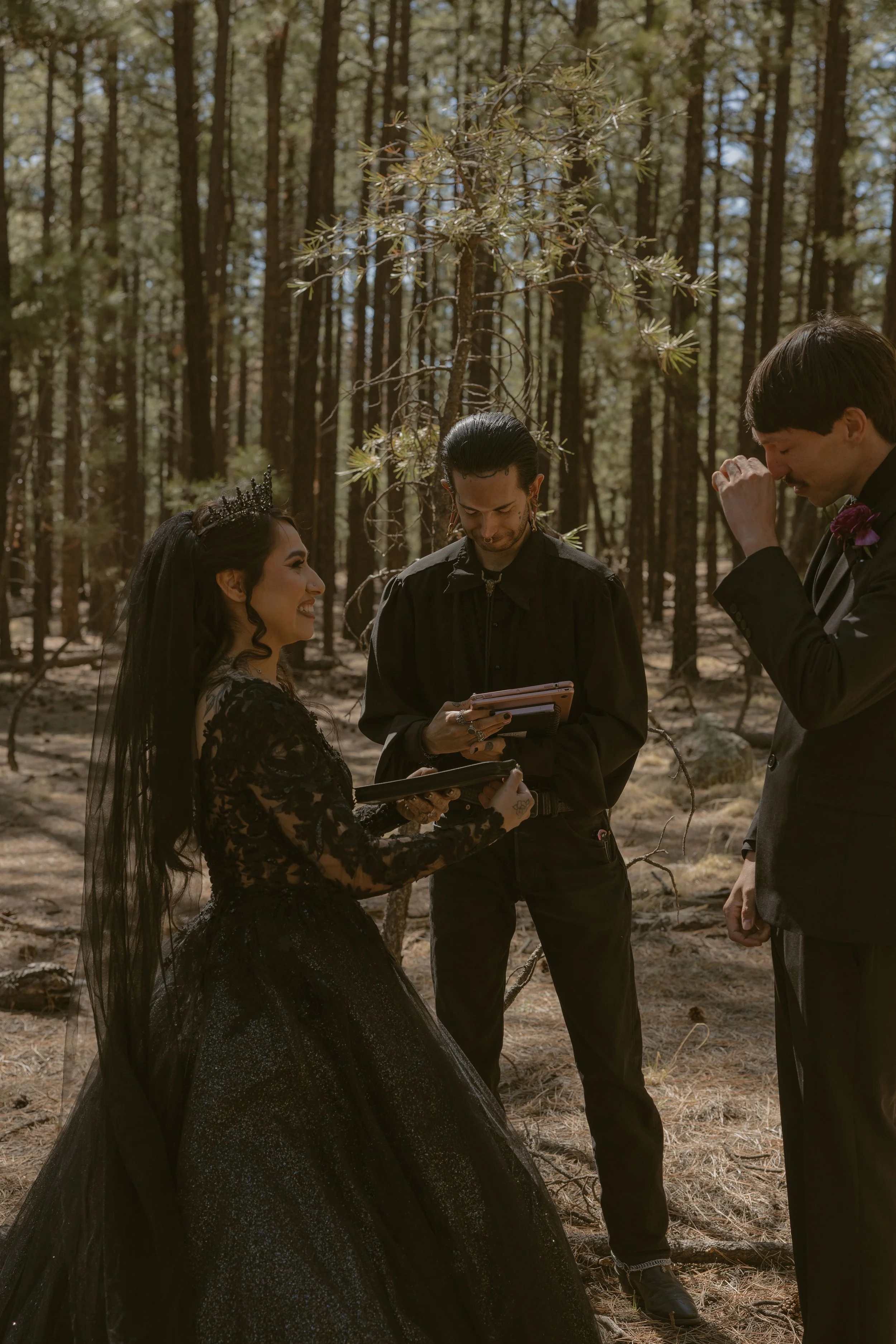 A woman in a black lace dress with a tiara is getting married in a forest, with two men exchanging vows or rings, one holding a clipboard and the other with a hand over his face in emotion, under tall trees.