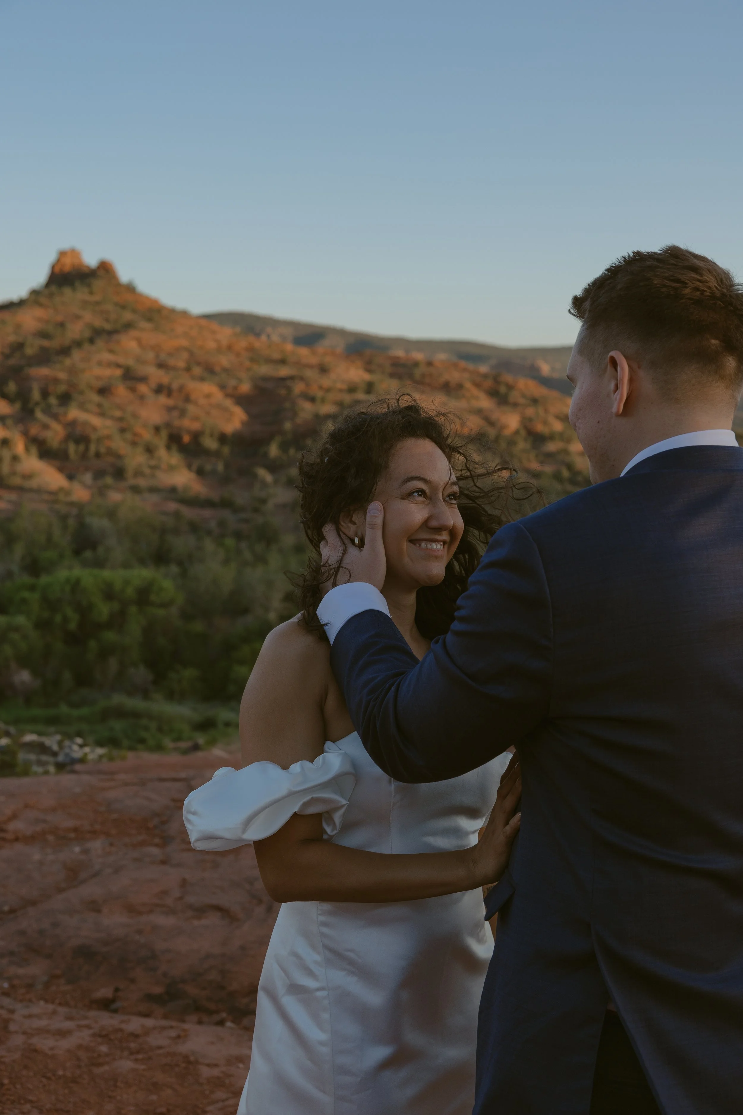 A woman in a white dress smiling at a man in a dark suit holding her face, outdoors with mountains and a blue sky in the background.