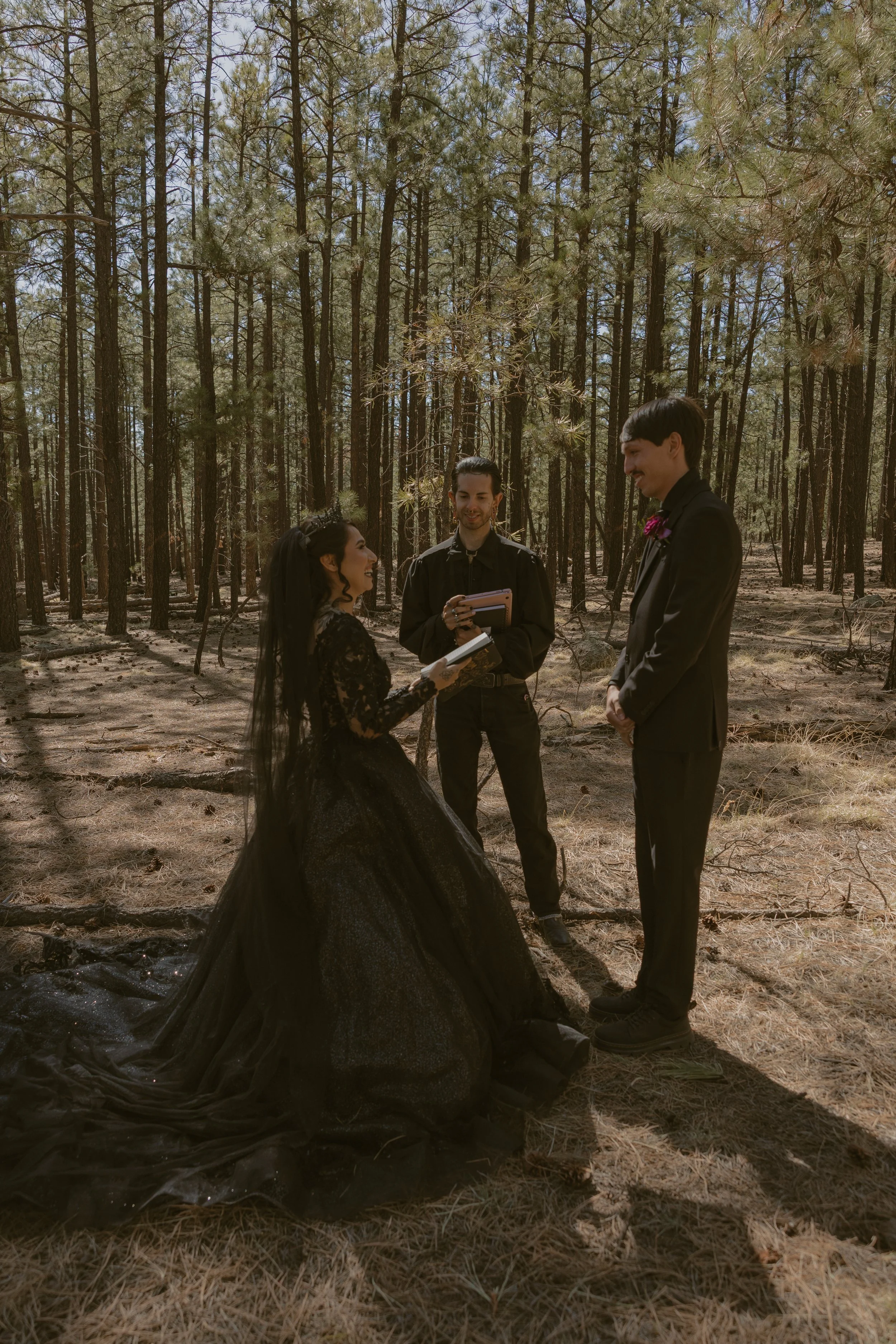 A wedding ceremony taking place outdoors in a forest with a bride and groom standing and exchanging vows, and two officiants taking notes.