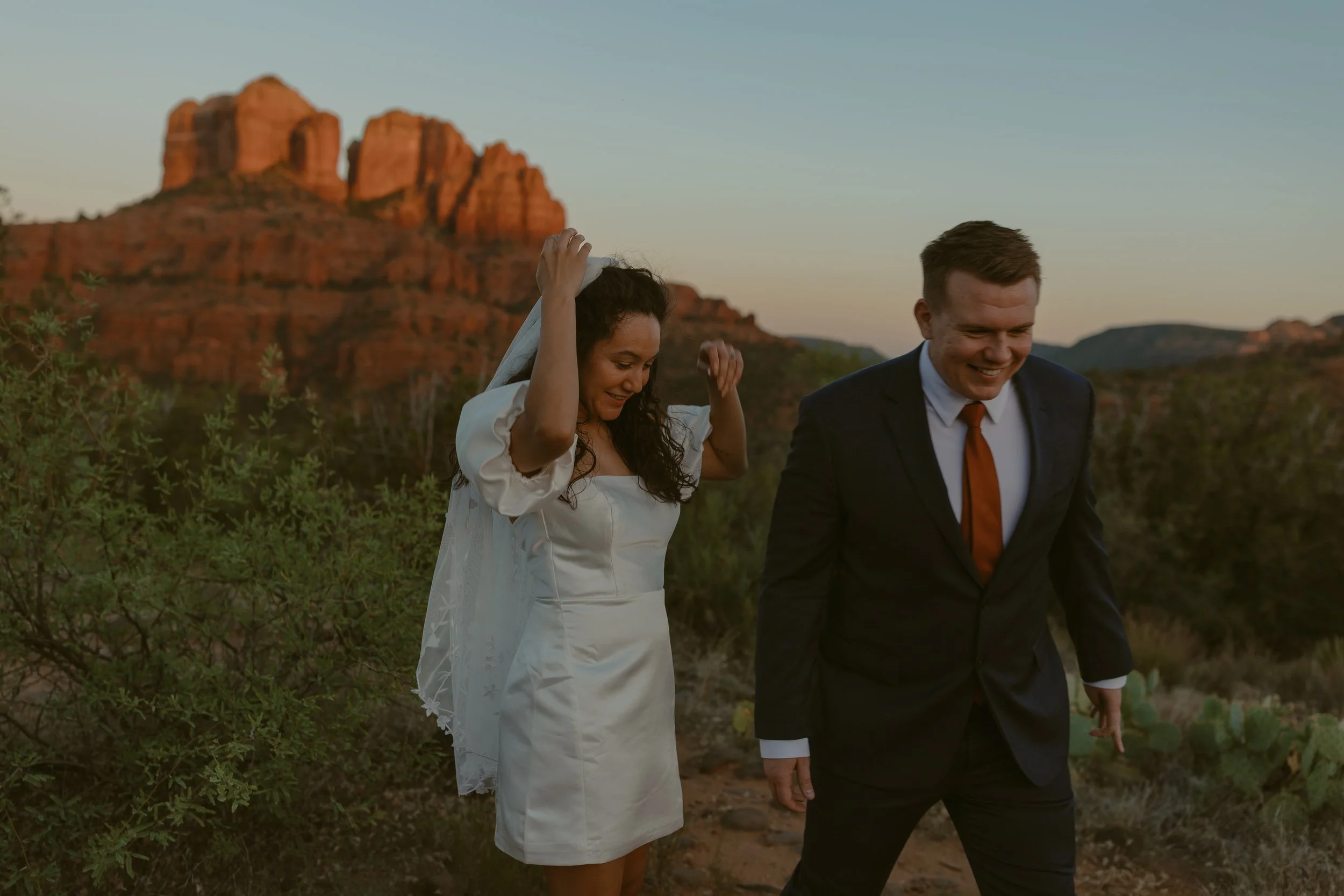A smiling woman in a white dress and a man in a suit walking outdoors at sunset in a desert landscape with red rock formations.