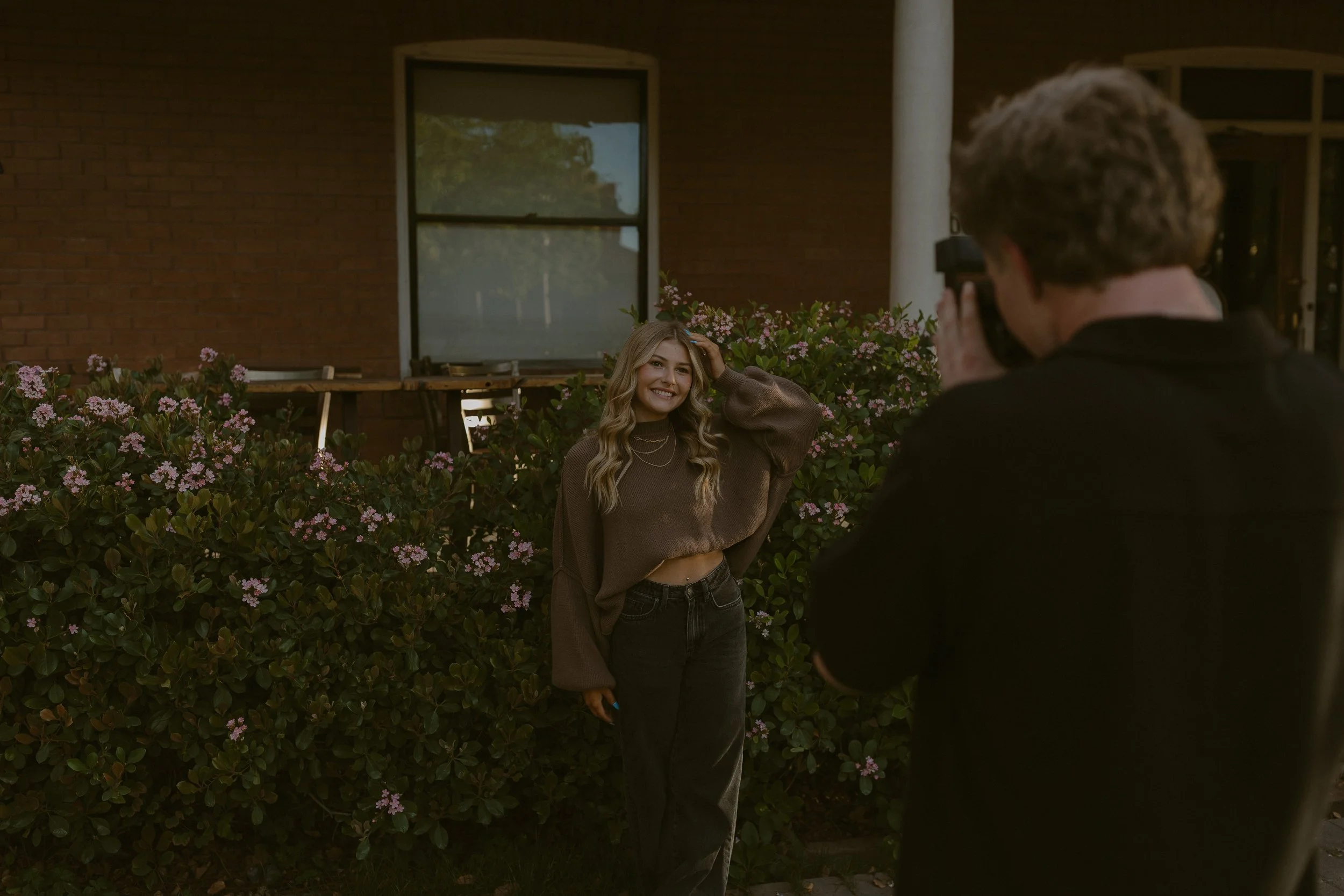 A person in a brown sweater posing for a photographer in front of a house with brick walls and flowering bushes.
