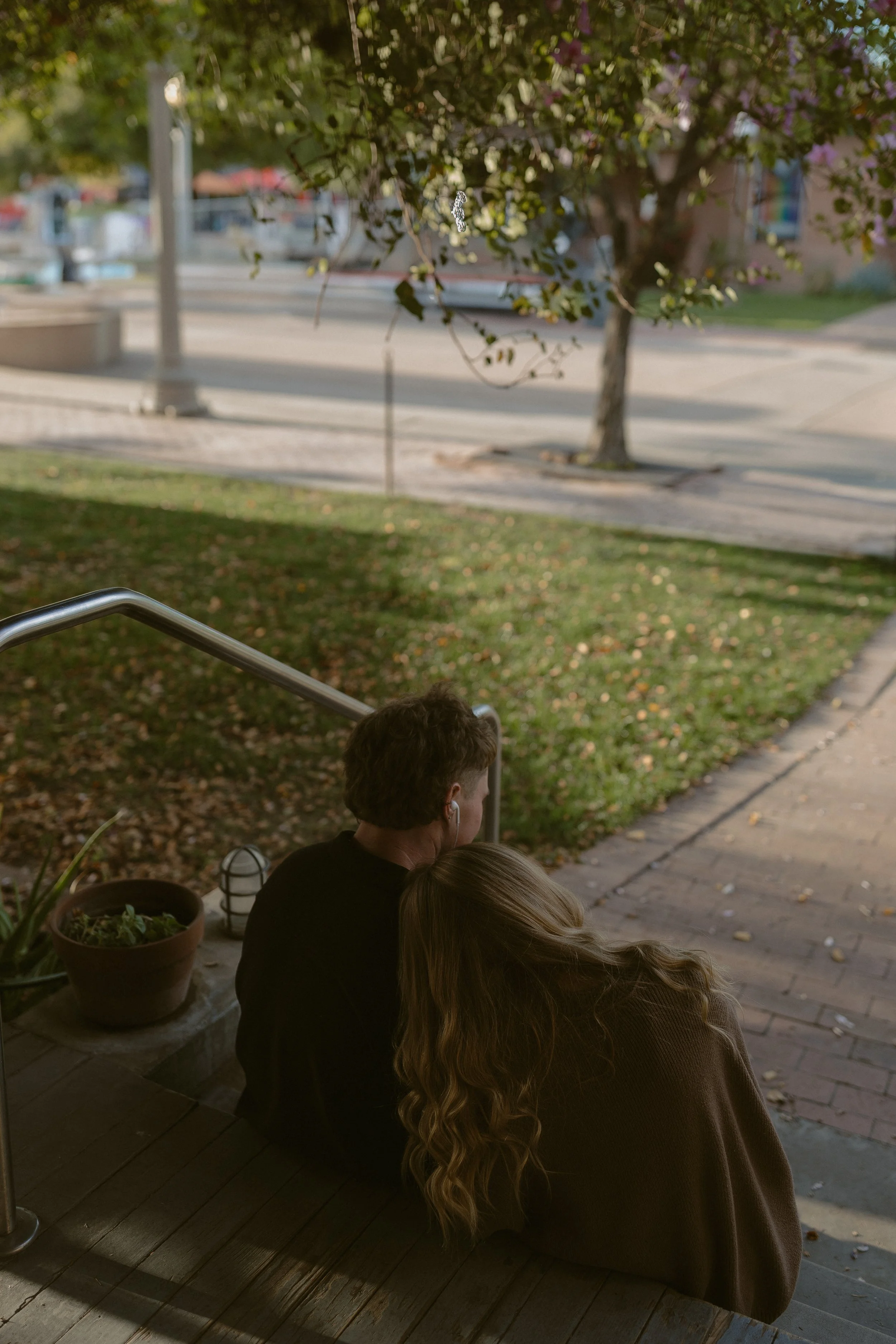 A couple sitting on outdoor steps by a garden, the woman resting her head on the man's shoulder, surrounded by trees and a paved area.