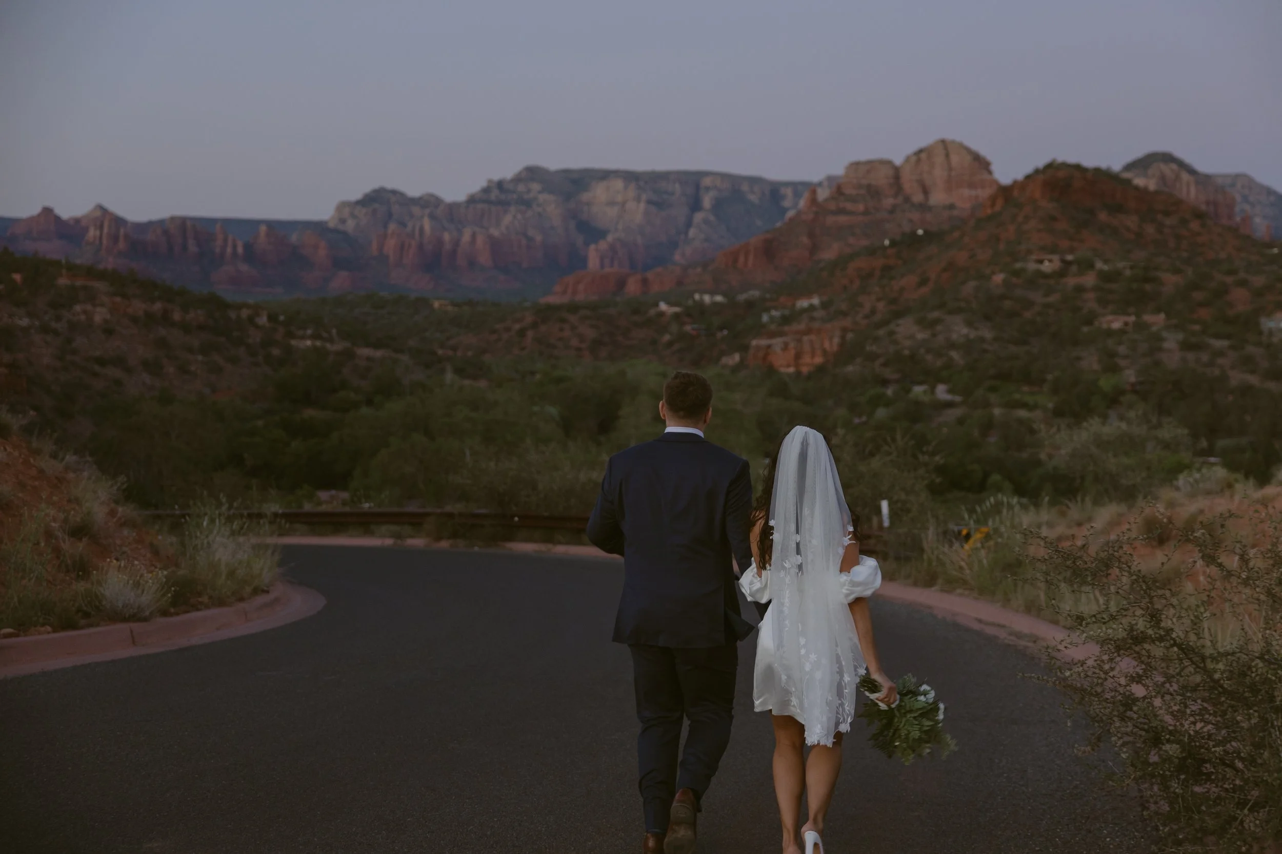 A newlywed couple, the groom in a black suit and the bride in a white dress with a veil, walking down a curved road in a desert landscape with red rock formations in the background during dusk.