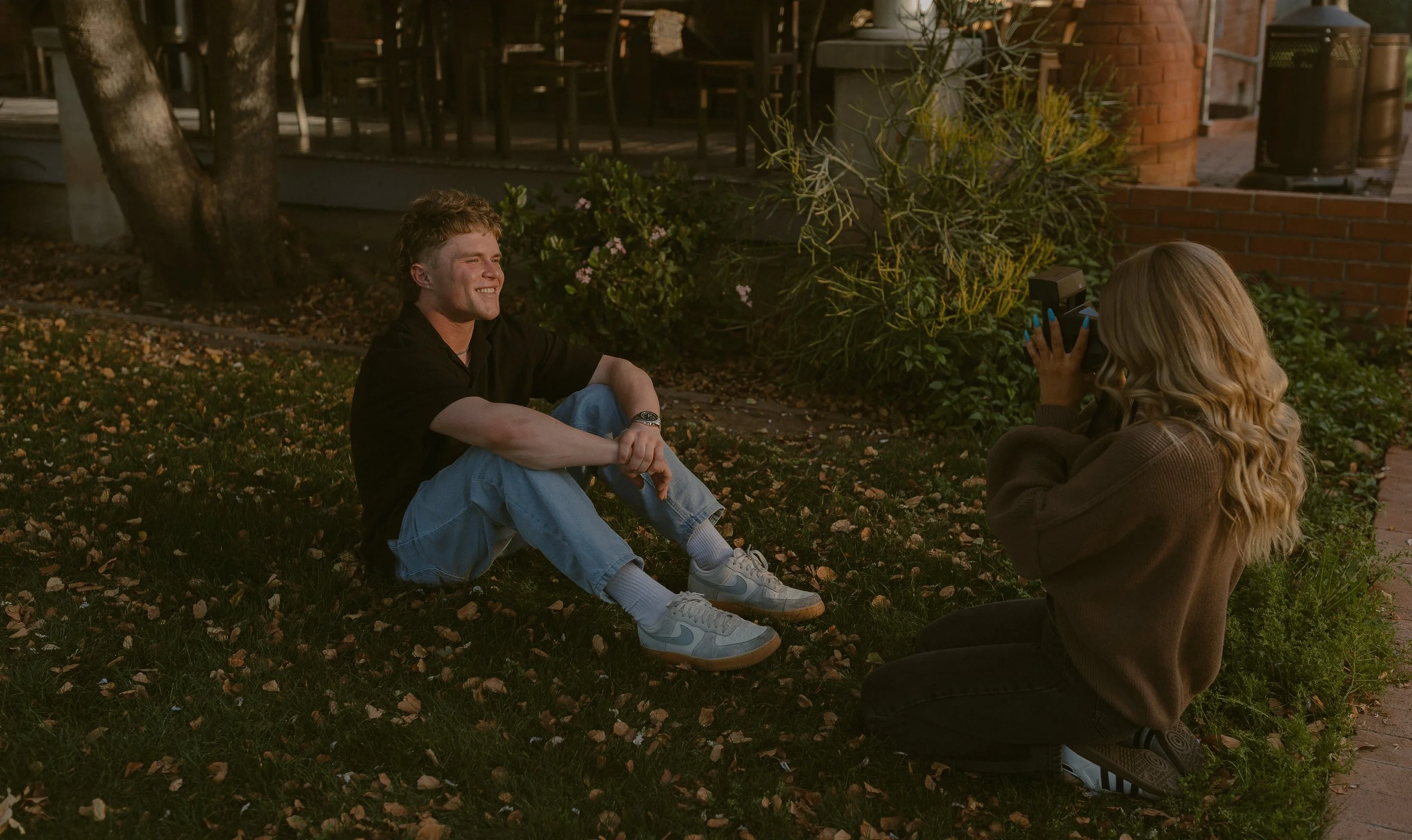 A person sitting on grass next to a tree, smiling as a woman with long blonde hair takes a photo of him using a vintage camera. Leaves are scattered on the ground, and a wooden structure is in the background.