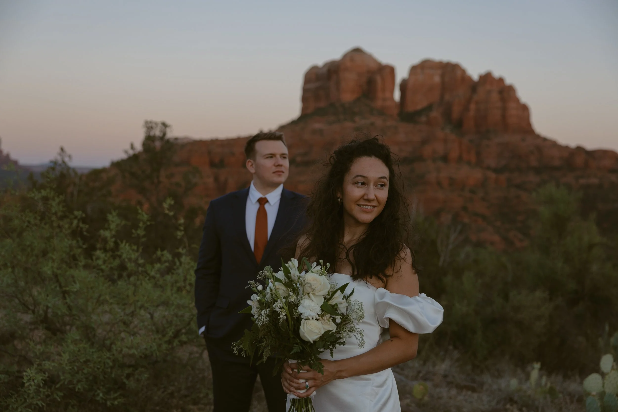 A couple on their wedding day standing outdoors with red rock formations and desert vegetation in the background at sunset.
