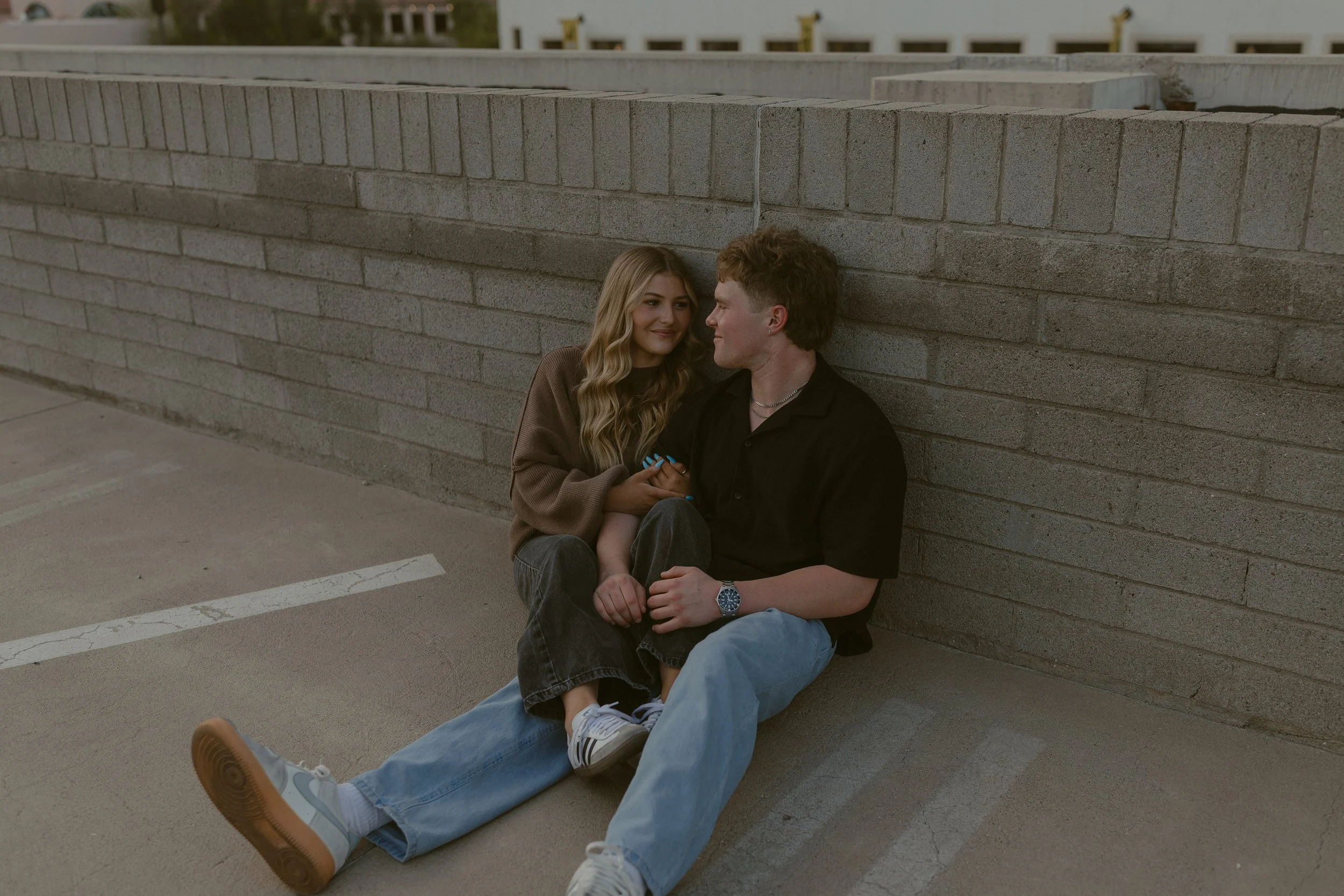 Young couple sitting on a rooftop, leaning against a brick wall, smiling at each other.