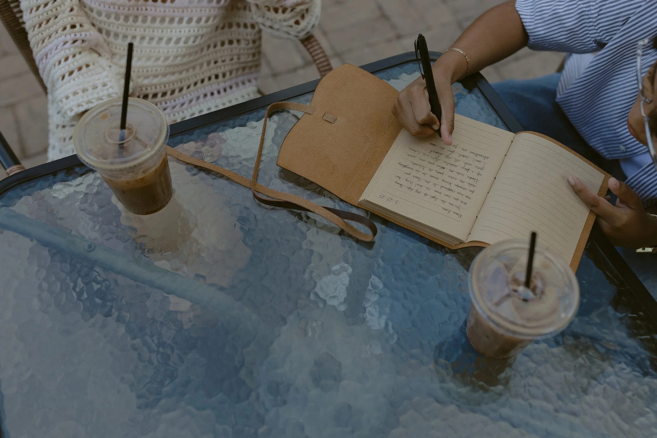 Two people sitting at a glass table with iced coffee drinks and an open notebook, one person is writing in the notebook.