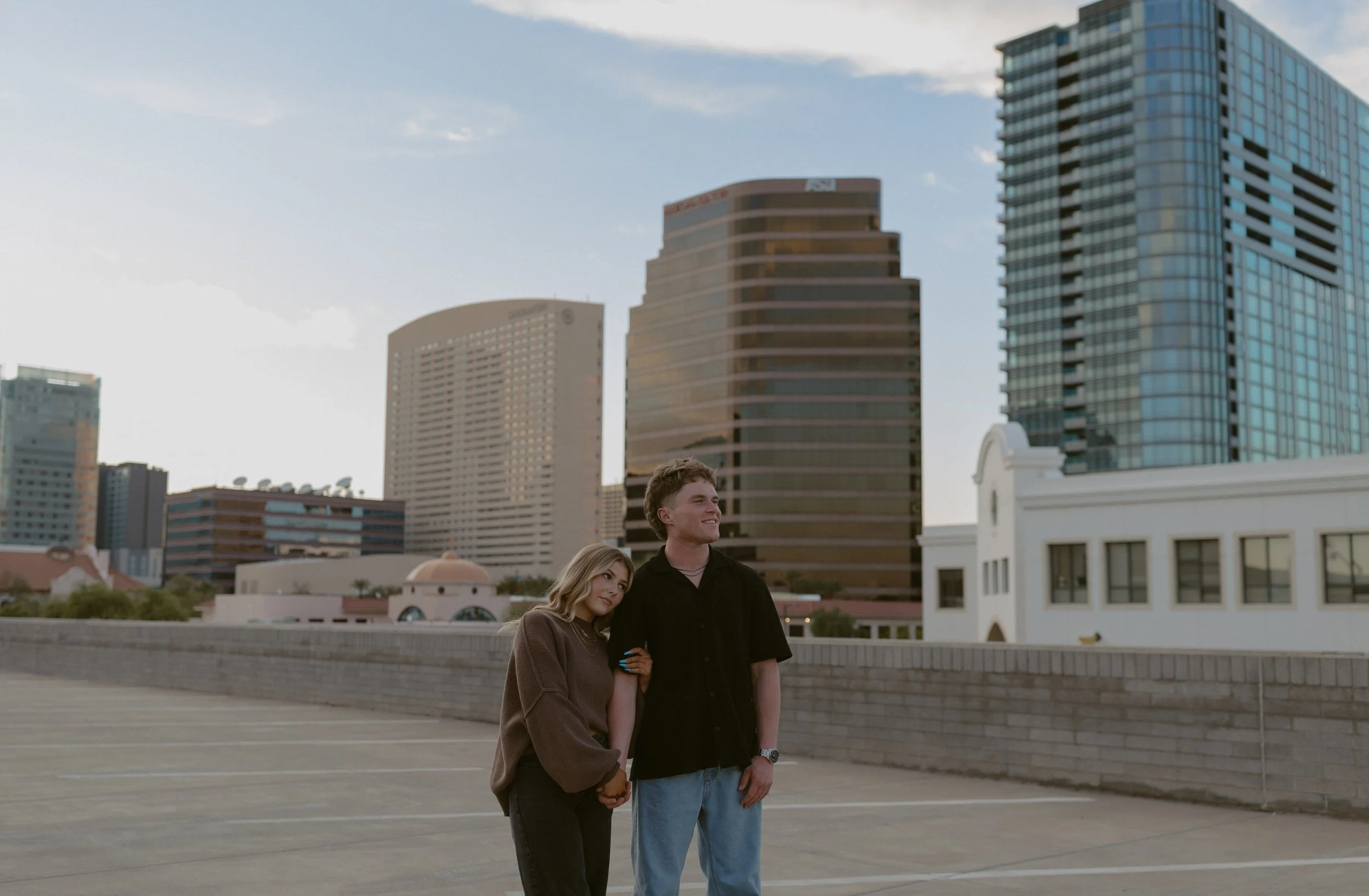 A couple stands on a rooftop parking lot with city buildings in the background. The woman leans on the man's shoulder, and they hold hands while looking into the distance. The scene is set against a clear blue sky.