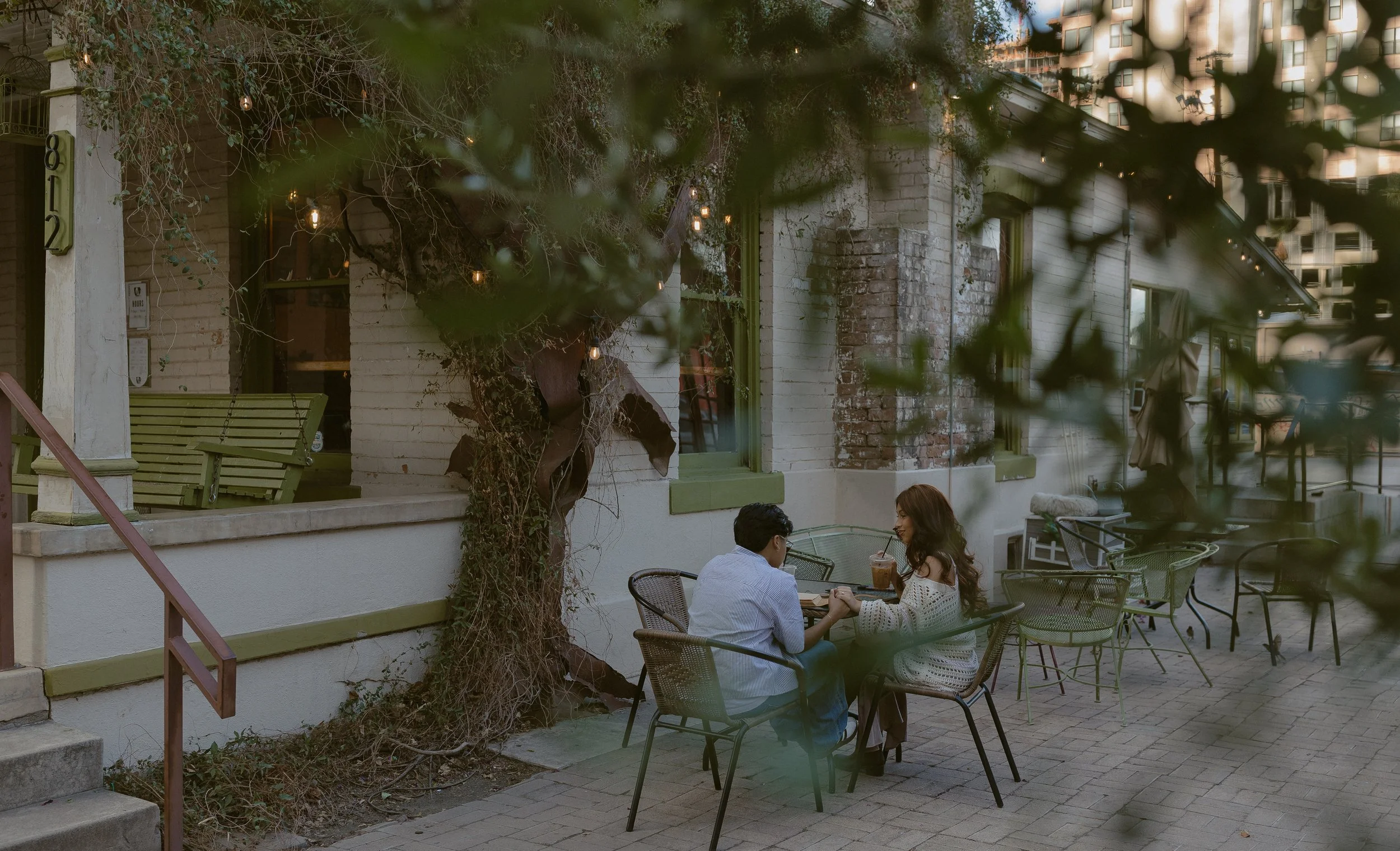 Two people sitting at an outdoor café table near a brick building with greenery and hanging lights.