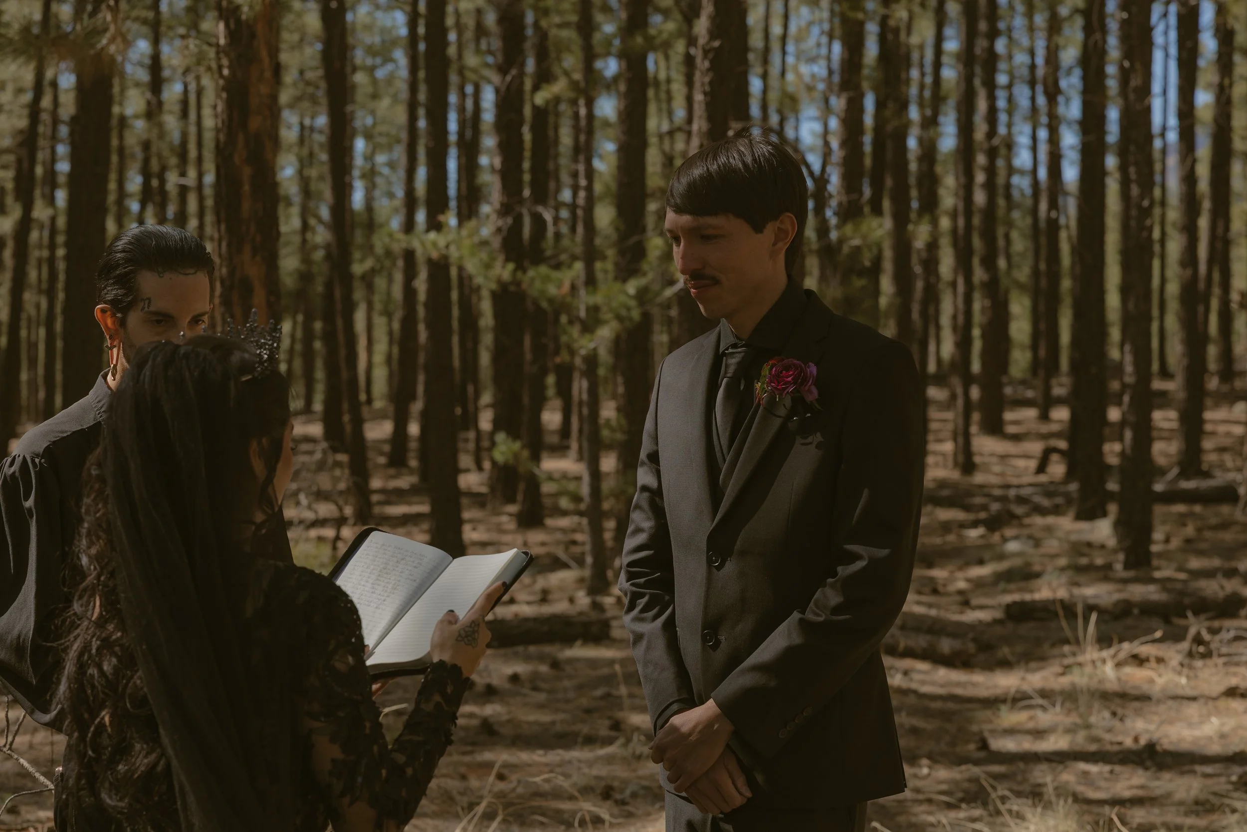 A wedding ceremony taking place in a forest, with a man in a gray suit standing in front of a woman reading from a book, and another woman assisting, during daytime.