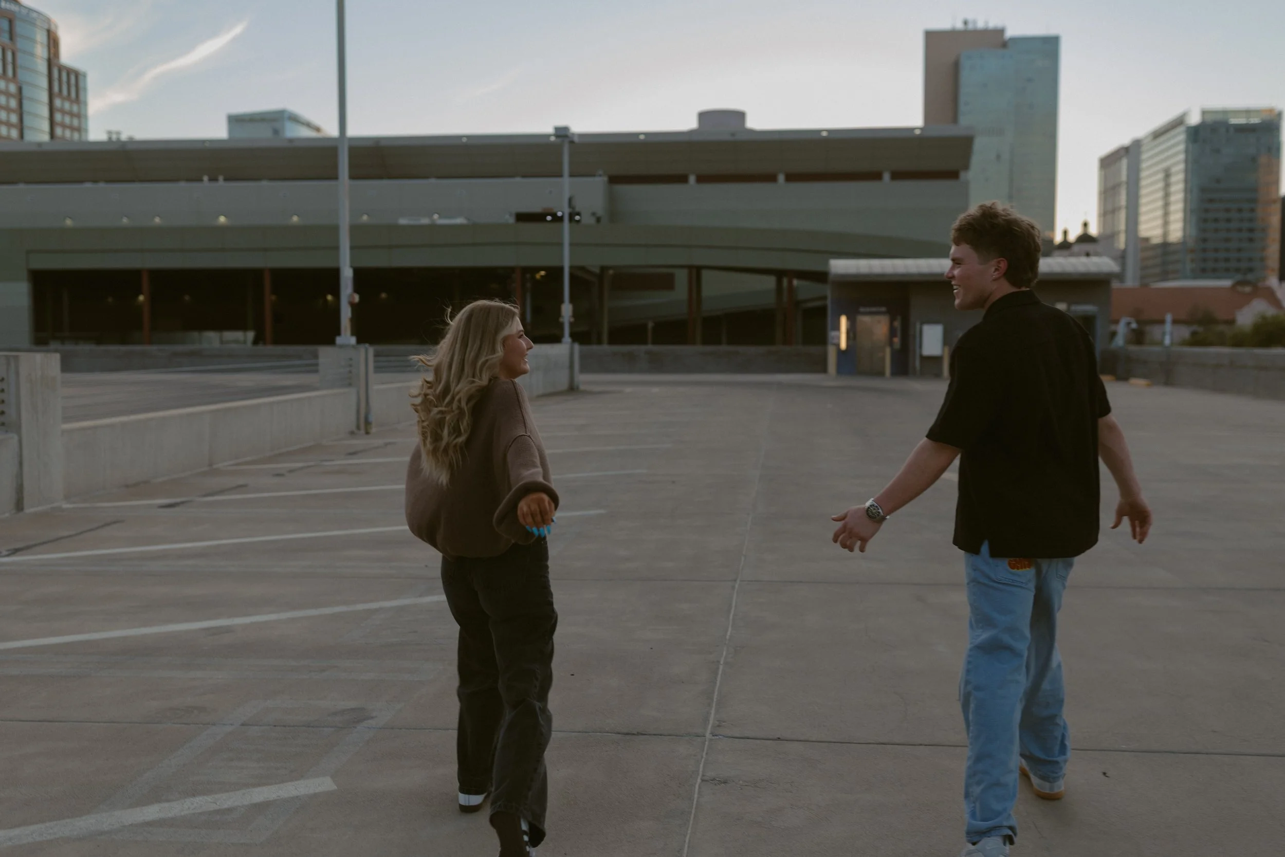 A young woman and man walking in an empty parking lot, holding hands, with city buildings in the background.
