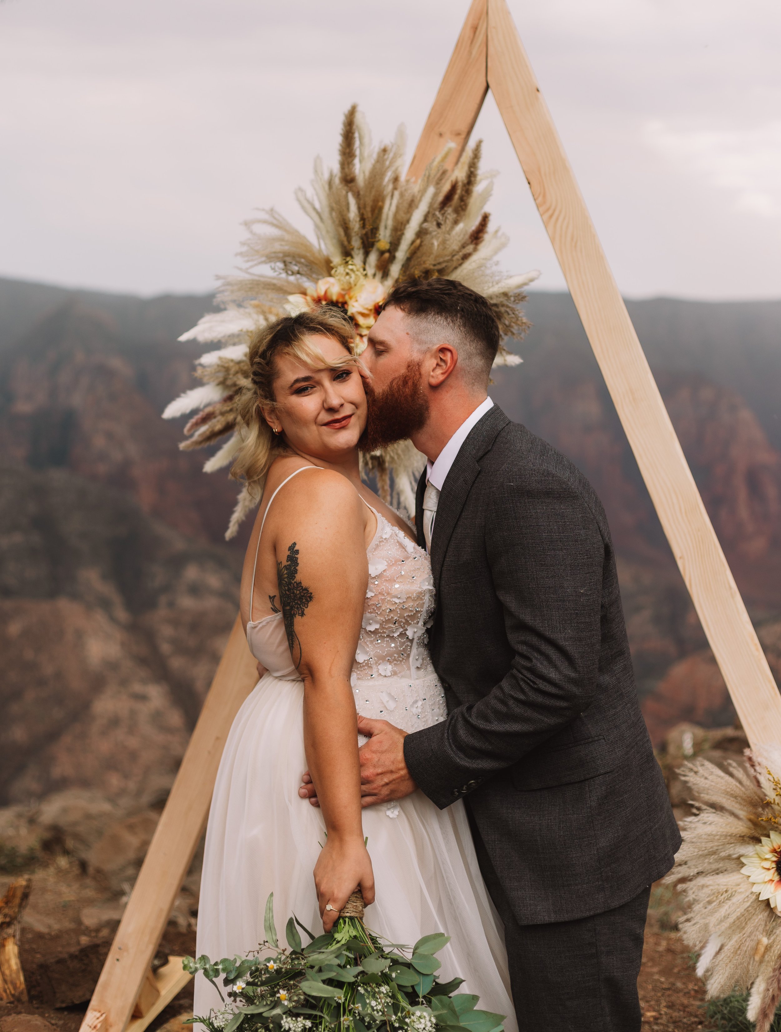 A couple in wedding attire poses outdoors near a triangular wooden arch adorned with pampas grass and flowers. The groom, in a gray suit, kisses the bride's forehead as she holds a bouquet of greenery and white flowers. A scenic mountain background c