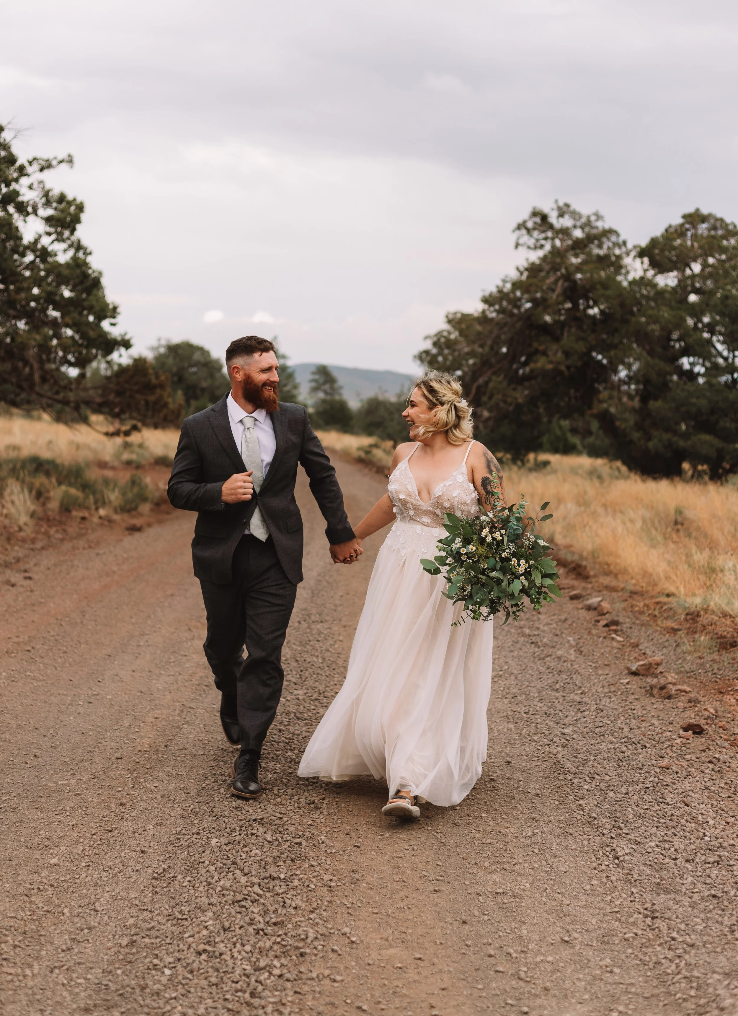 A bride in a white dress and groom in a suit holding hands while walking on a gravel road, surrounded by trees and grass, with cloudy skies above.