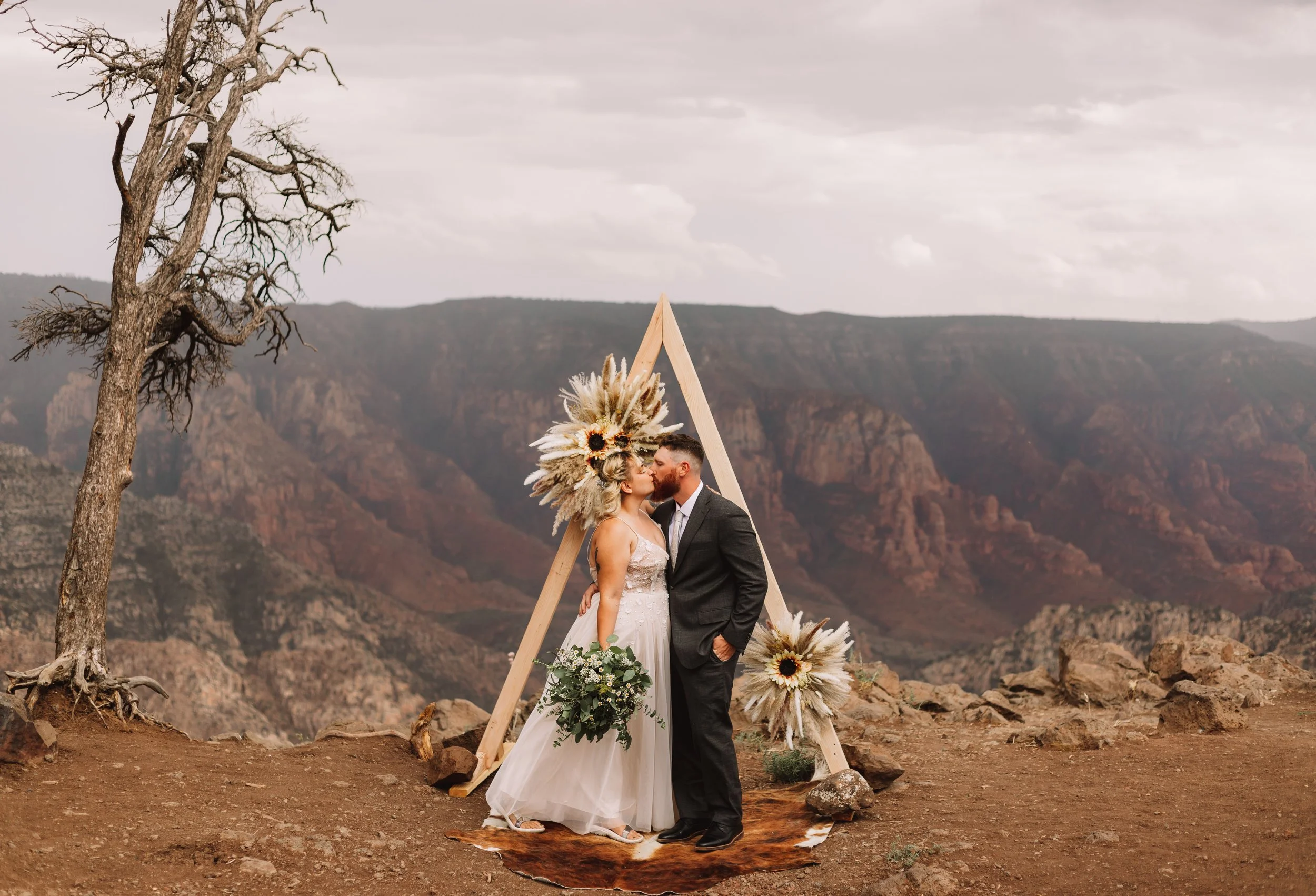 A couple in wedding attire kissing in front of a triangular wedding arch decorated with pampas grass and sunflowers, set on a rocky mountain overlook with an expansive canyon view.