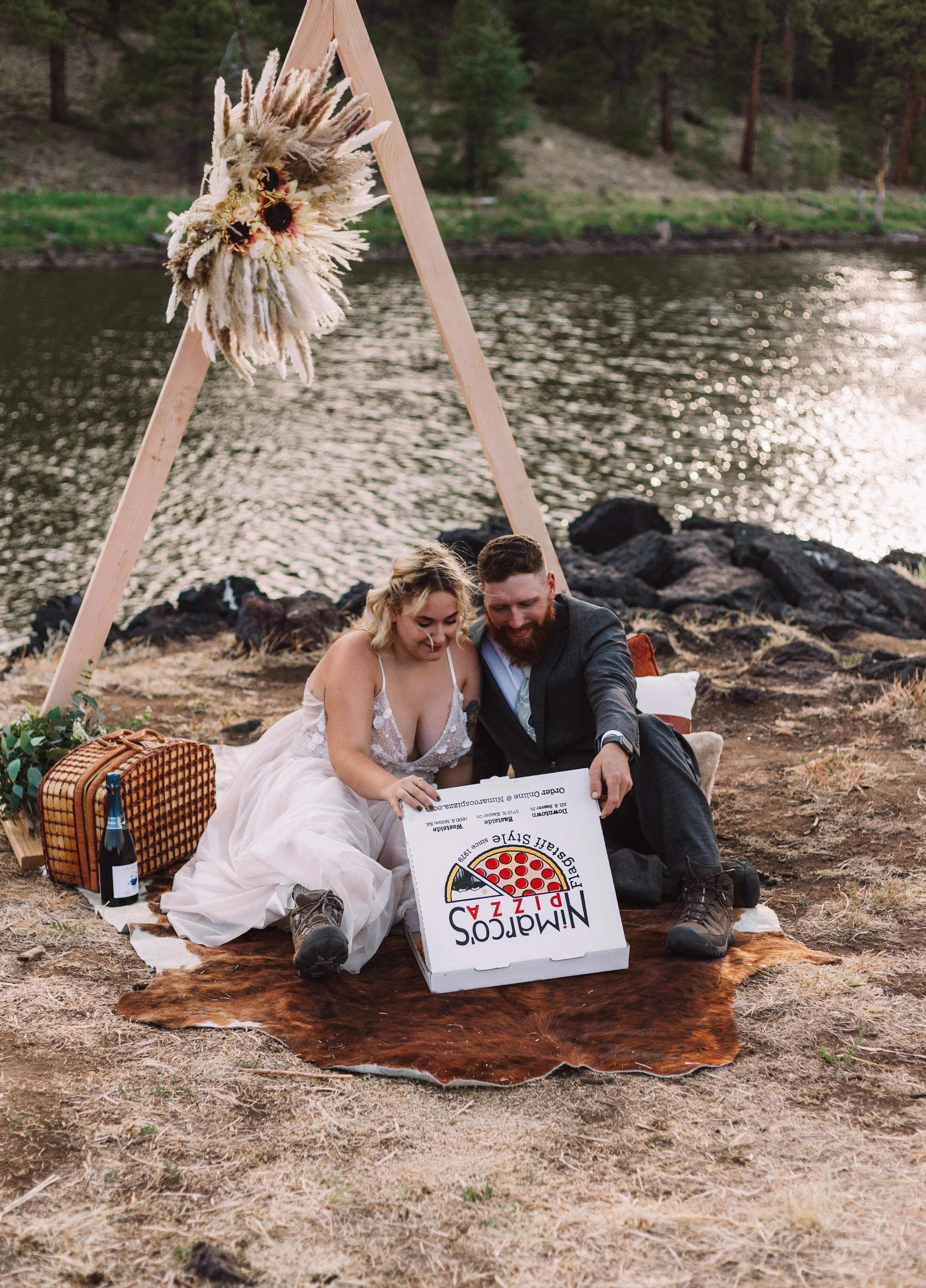 A couple sits on a blanket near a riverside, with a triangular wooden arch decorated with flowers behind them. They are looking at a pizza box labeled "Nimacos Pizza." A picnic basket and a bottle of sparkling wine are on the ground next to them. The