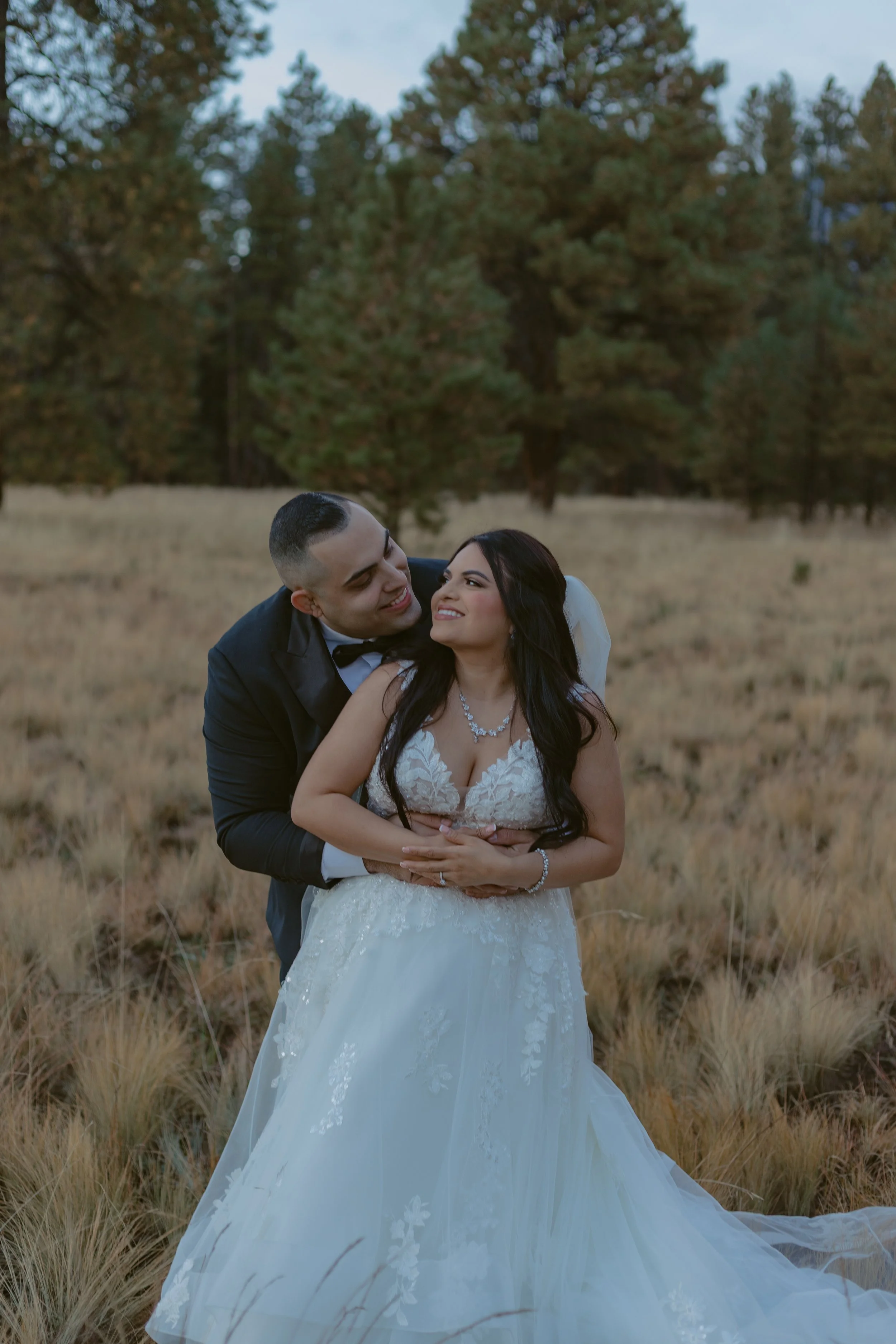 A newlywed couple sharing a moment in a field with trees in the background, the groom dressed in a black tuxedo and the bride in a white lace wedding gown.