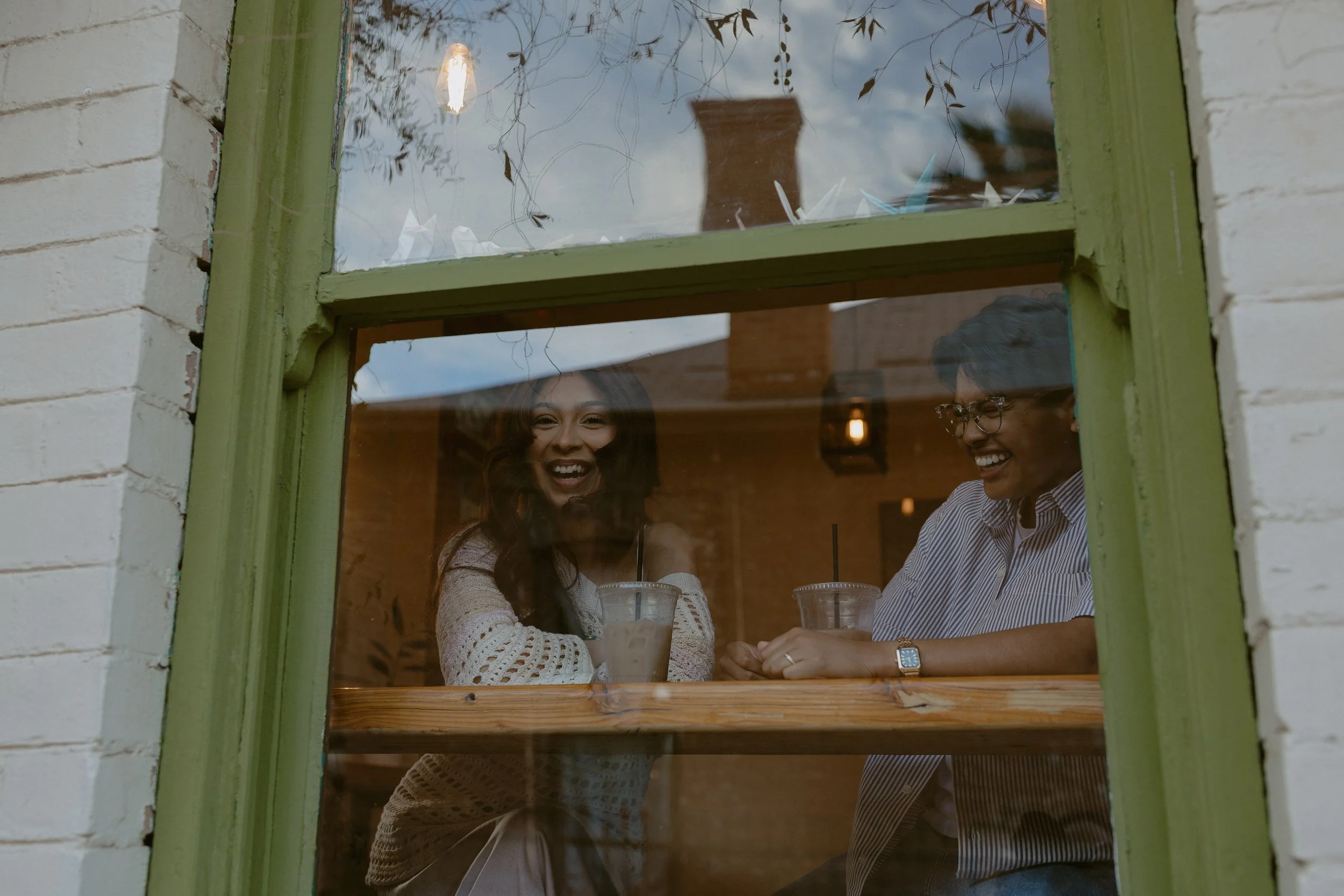 Two women sitting at a café window, laughing and enjoying drinks, as seen through a green-framed window in a cozy indoor setting.