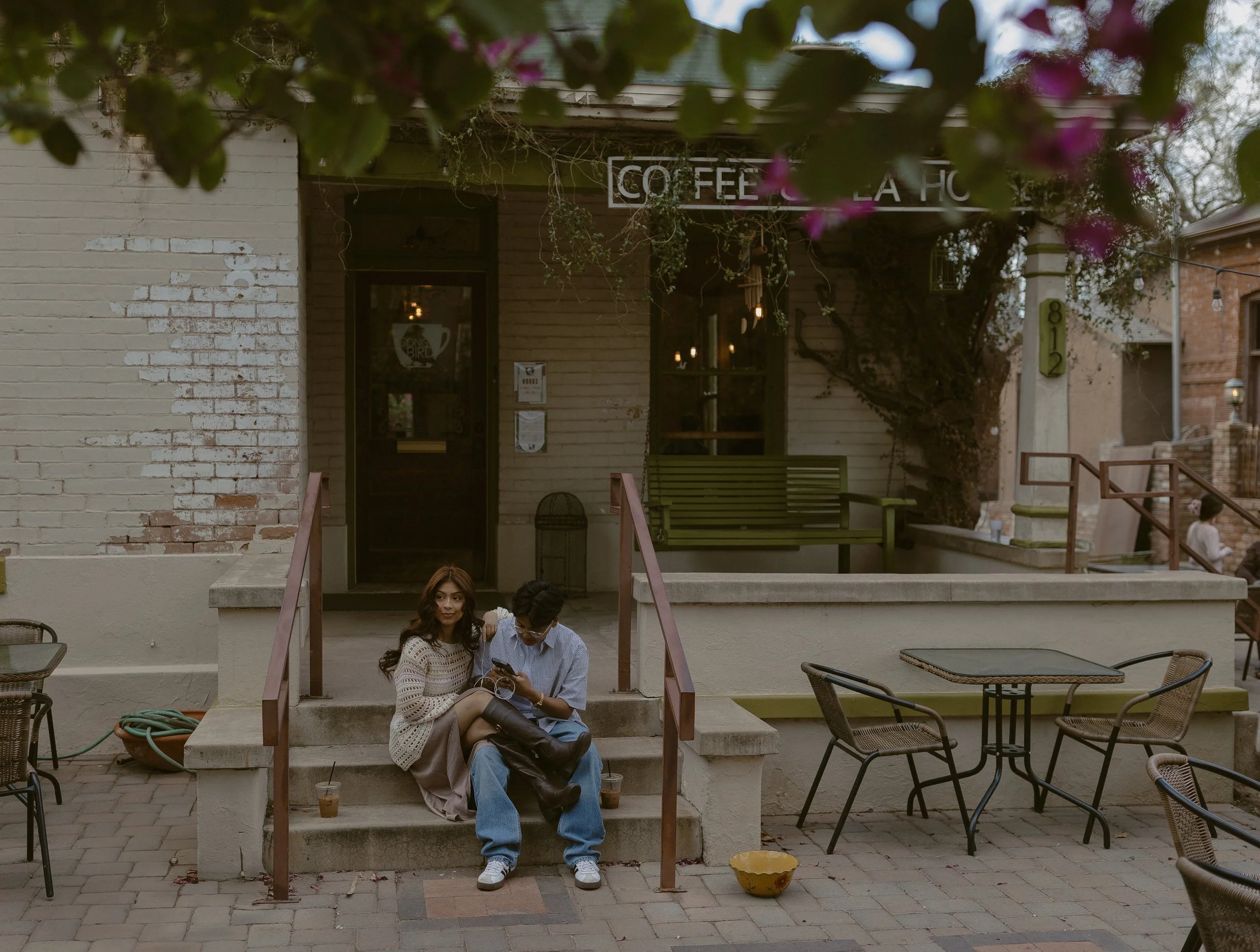 Two people sitting on the steps of a rustic coffee shop with a sign above the door. Outdoor seating and plants are visible, and there is a warm, relaxed atmosphere.