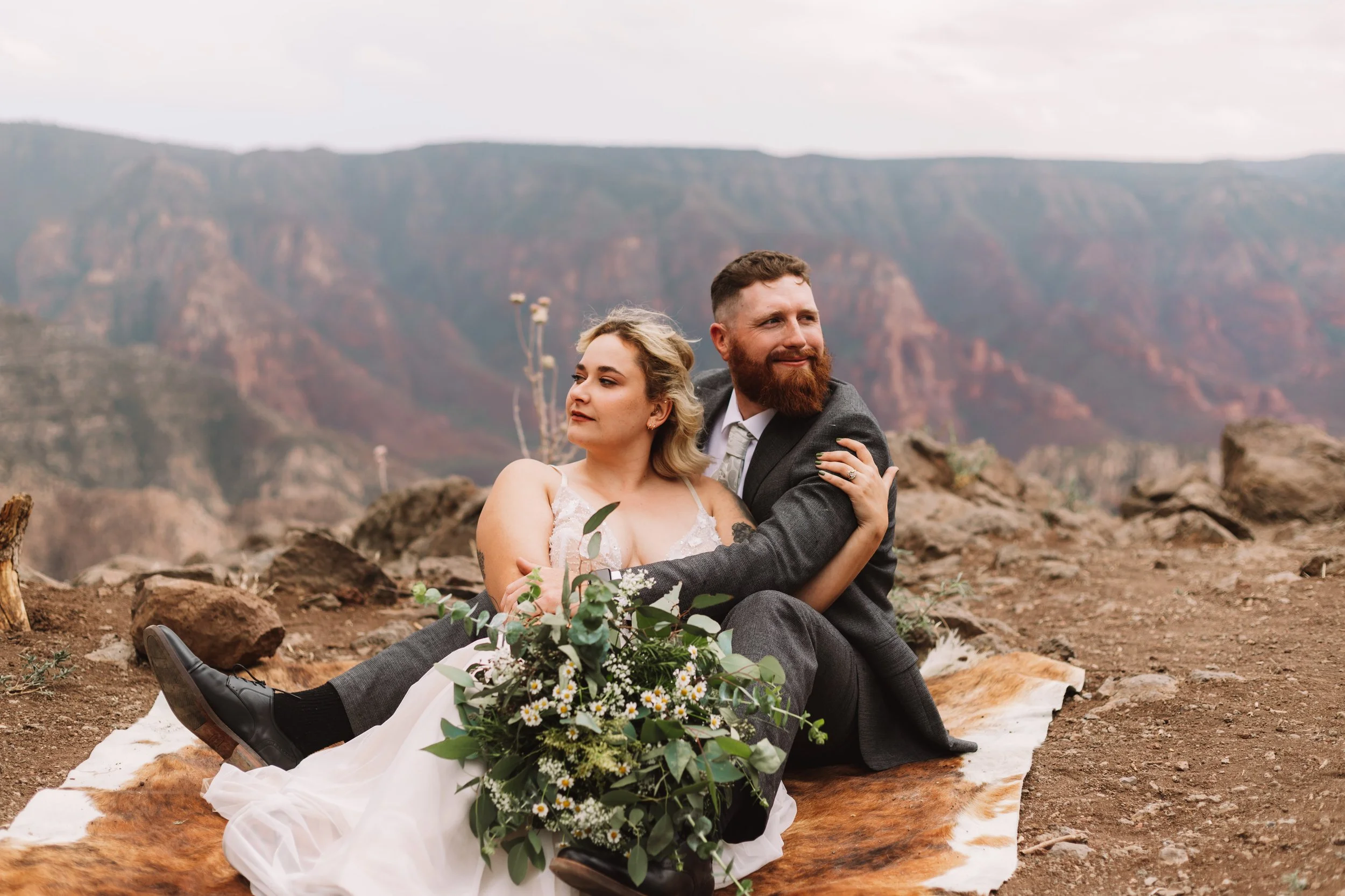Couple in wedding attire sitting on a blanket on a rocky cliff with mountainous landscape.