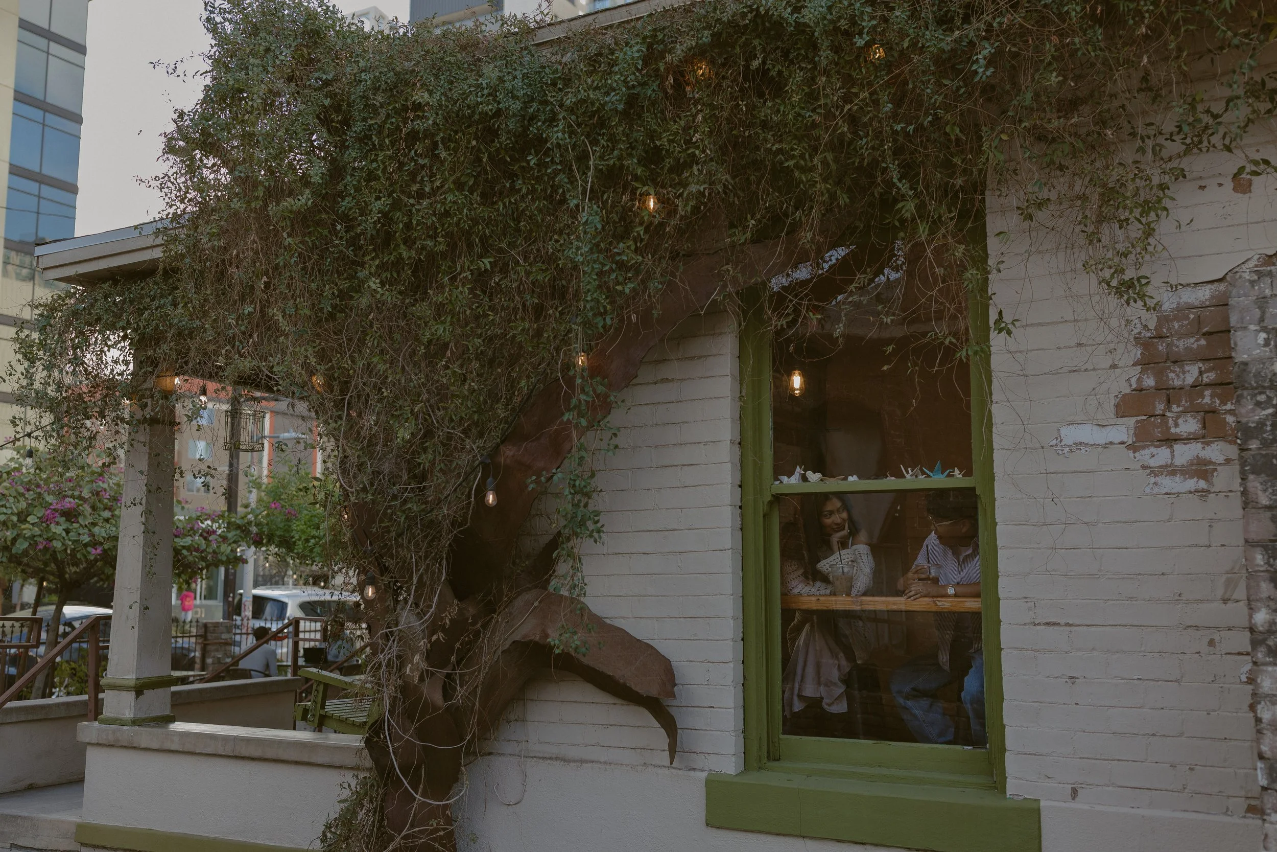 People sitting inside a cafe, visible through a window surrounded by a brick wall and overgrown vines with hanging lights outside.