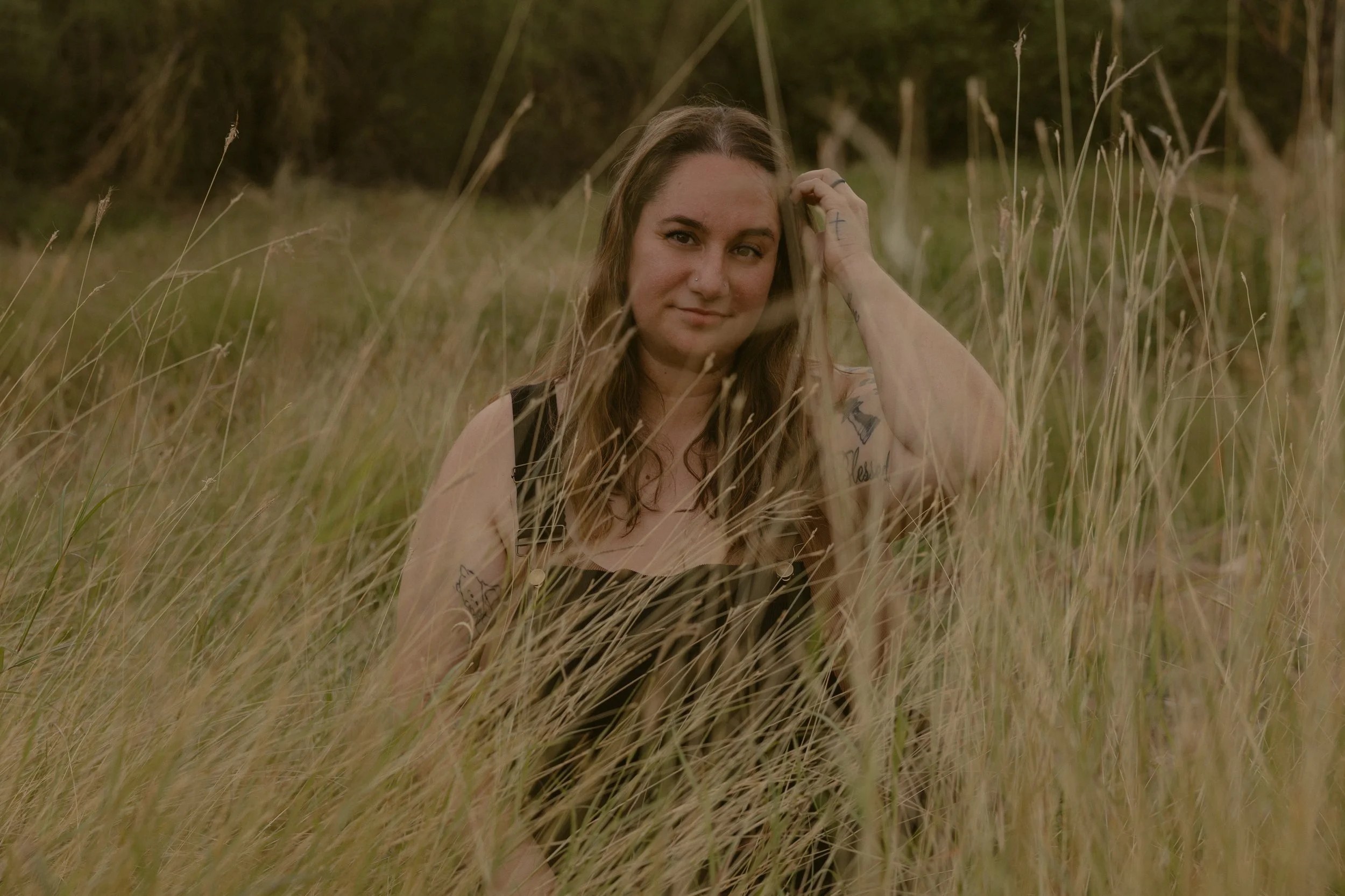 A woman with long wavy hair and tattoos on her arm sitting in tall grass in a natural outdoor setting.
