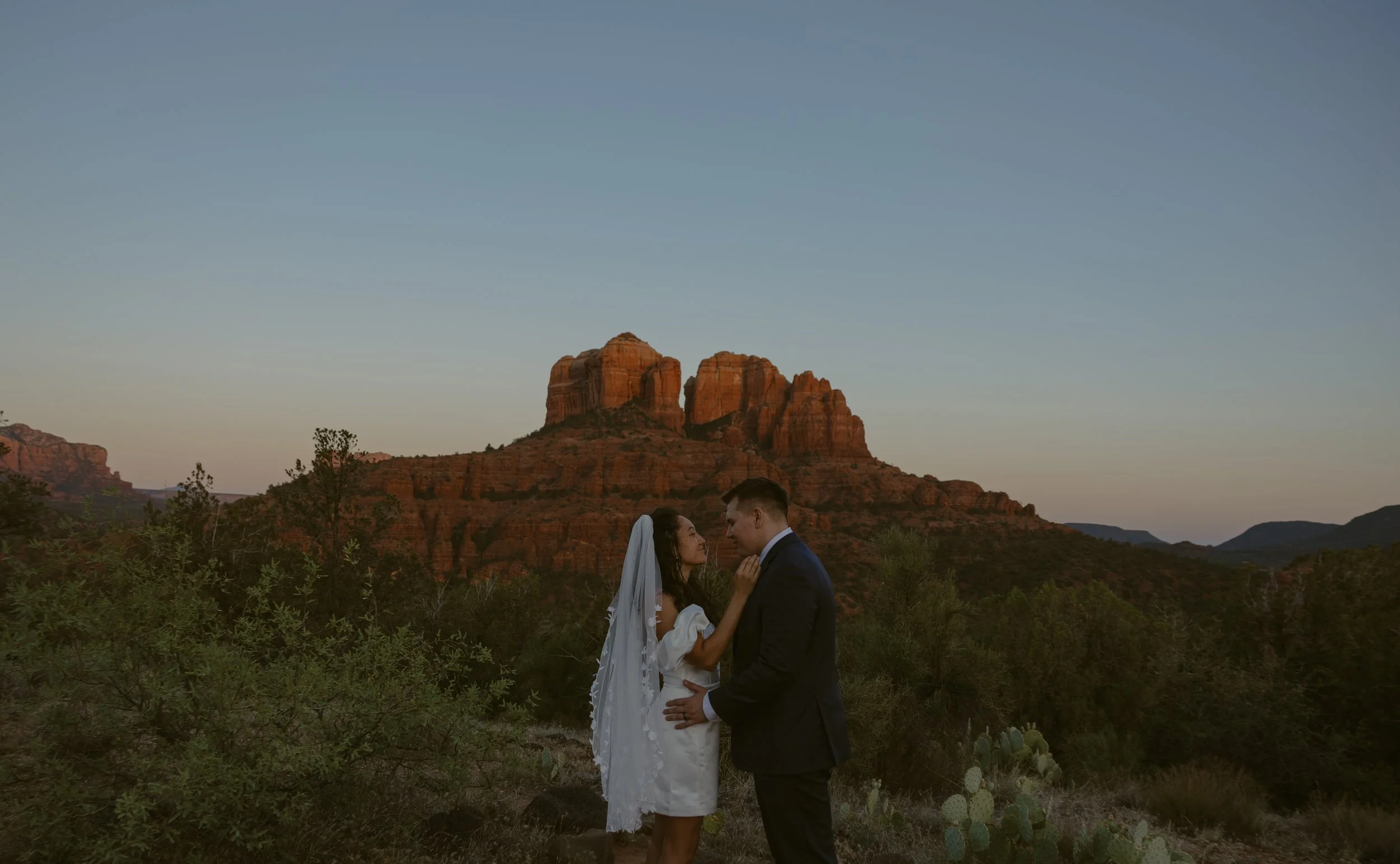 A bride and groom stand facing each other in a desert landscape at sunset, with large red rock formations in the background.