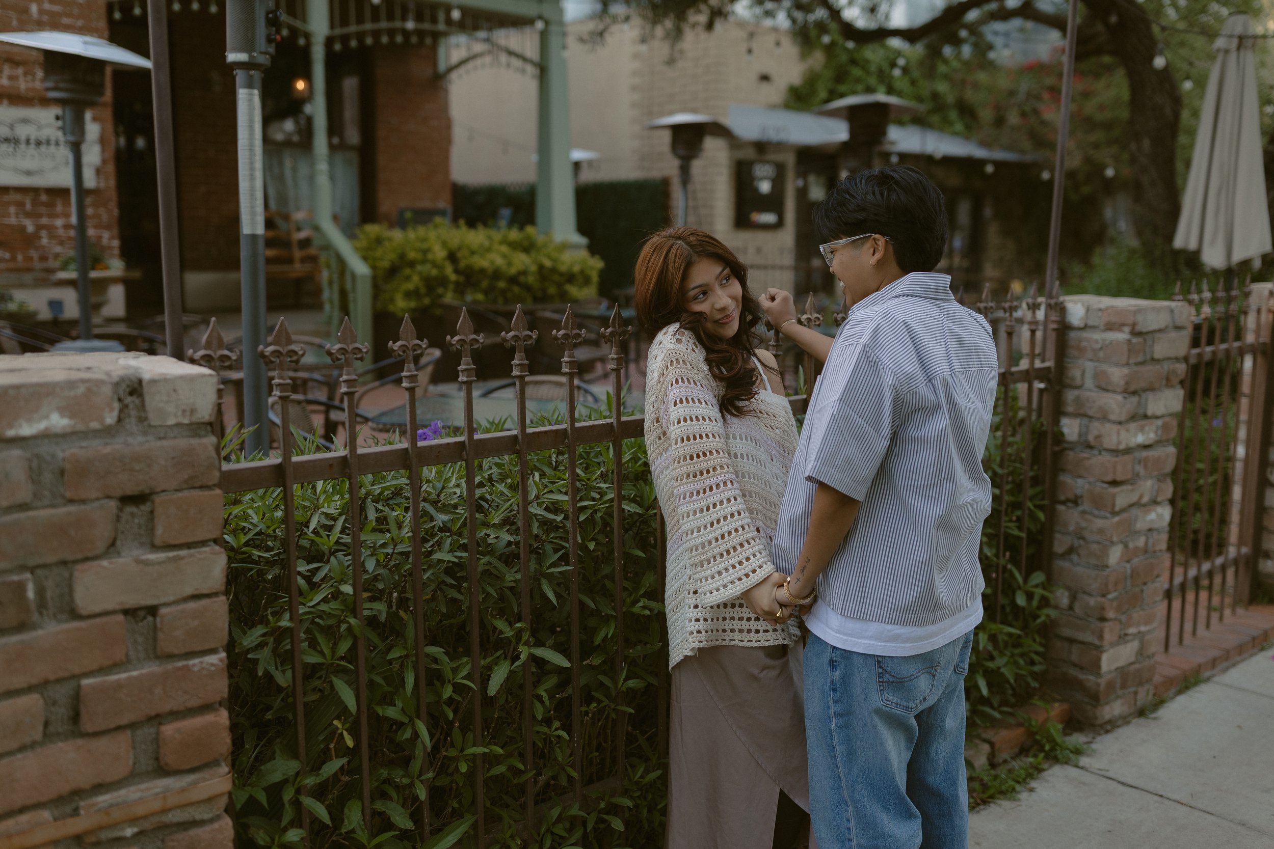 A young couple holding hands and looking at each other with smiles, standing in front of a decorative iron fence with green bushes behind it. The woman has wavy hair and is wearing a white crochet sweater and beige skirt. The man has dark hair, glass