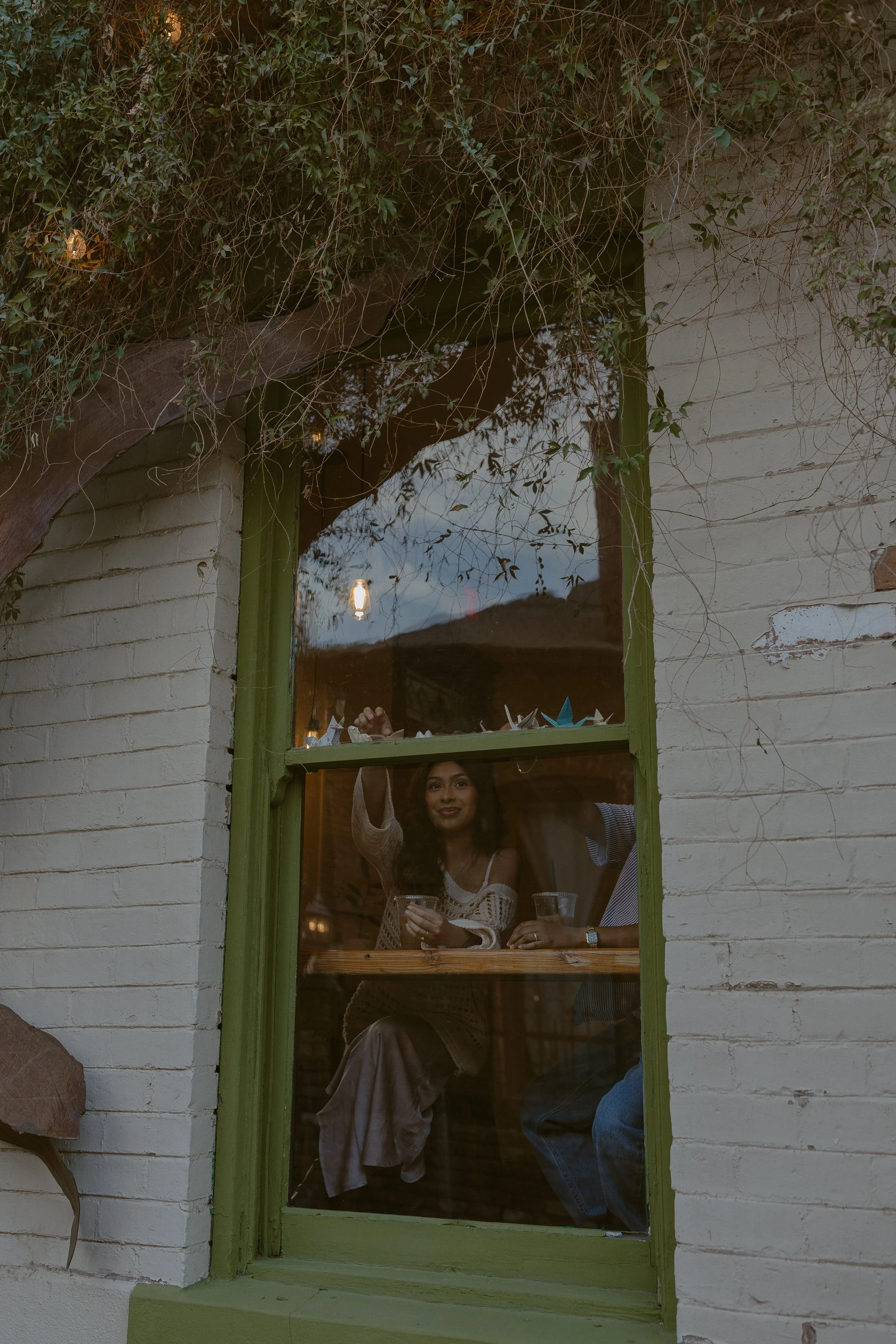Two people sitting inside a cafe, seen through a green-windowed exterior with vines and brick walls. One raises their hand, the other holds a drink.