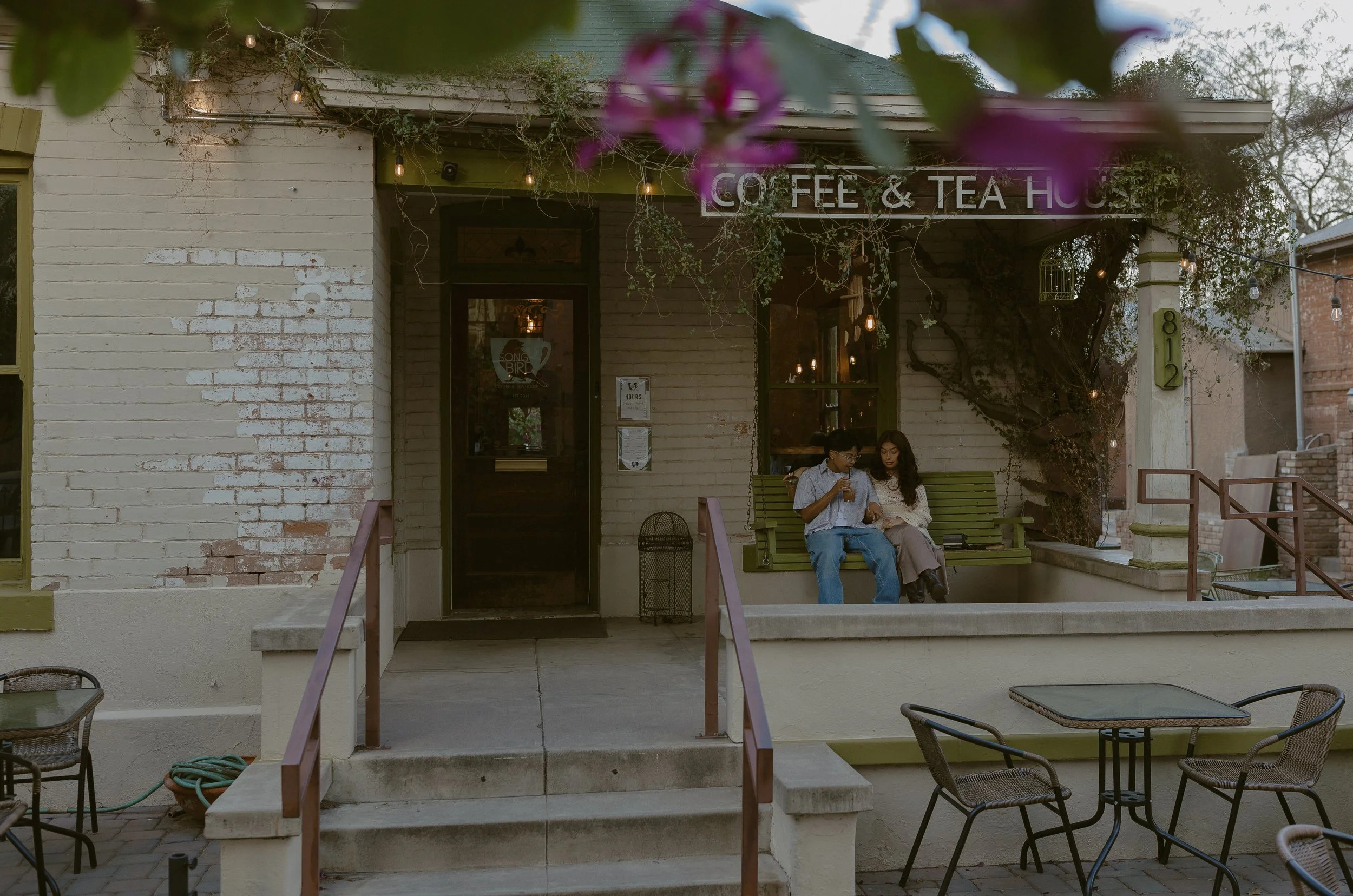 Exterior view of a coffee and tea house with a green bench where two people are sitting, one holding a drink. The building has a white brick facade with some exposed brick patches, and a purple sign reading 'Coffee & Tea House'. There are outdoor tab
