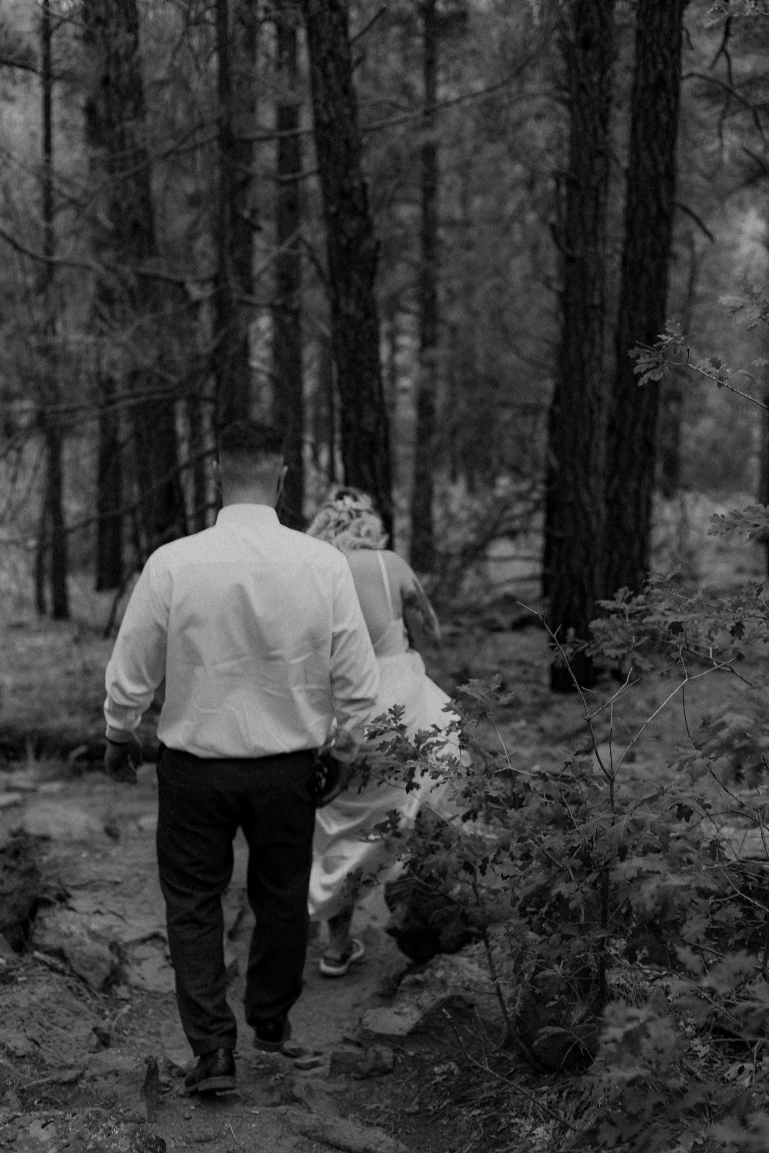 A man and a woman walking together in a forest, viewed from behind. The man is wearing a white shirt and dark pants, and the woman is dressed in a gown. The scene is in black and white.