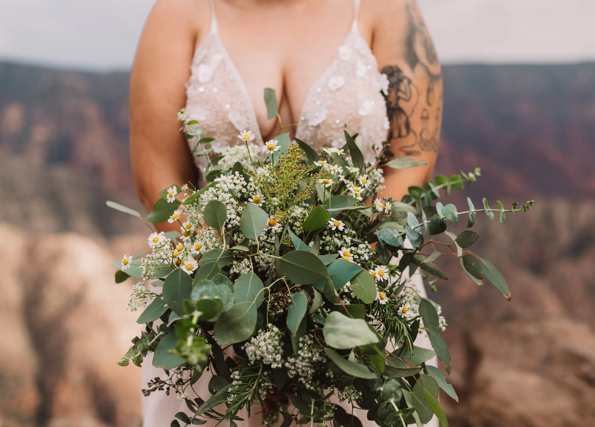 Bride holding a bouquet with greenery and white flowers, wearing a wedding dress with a visible tattoo on her arm.