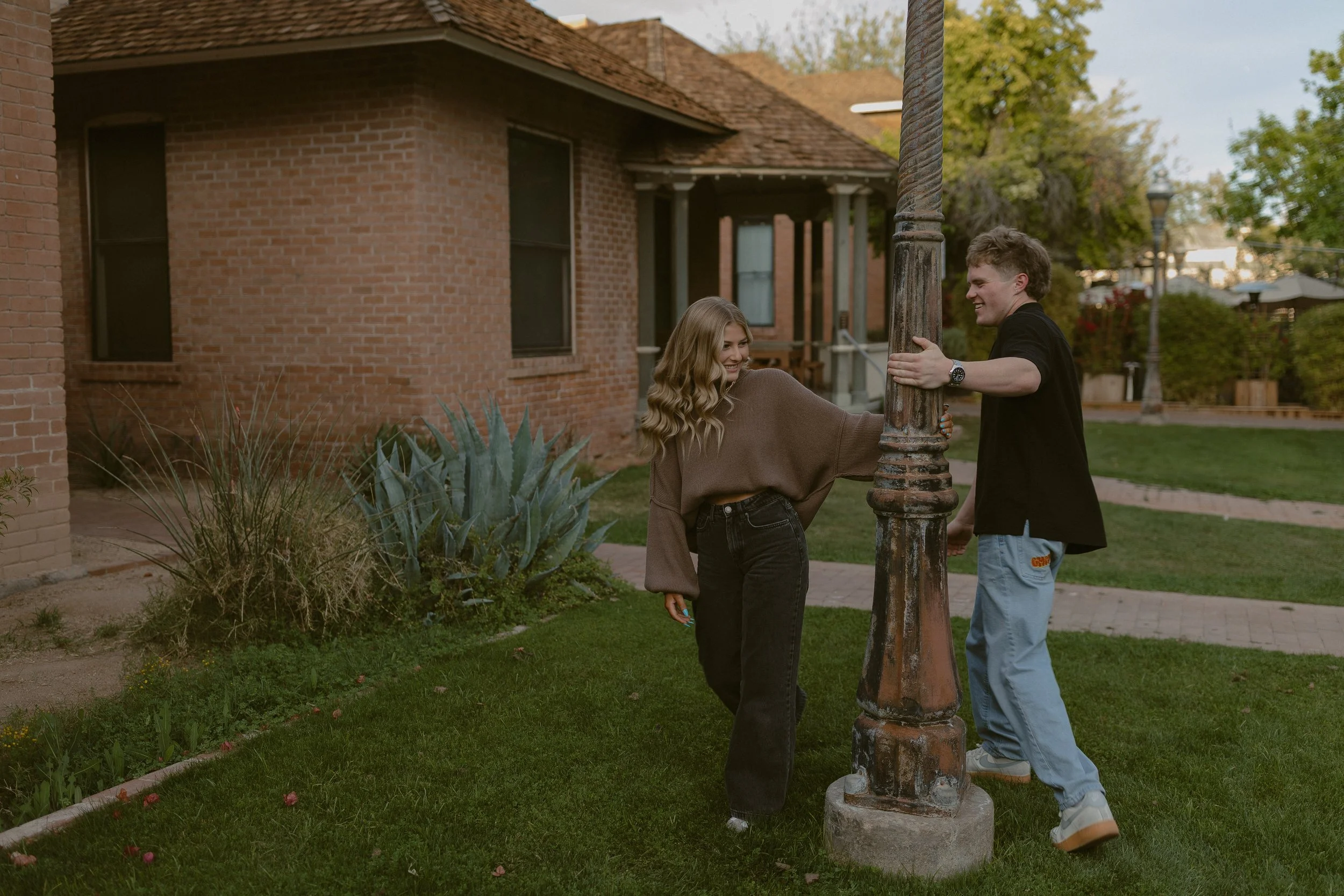 Two people smiling and interacting around a lamppost in a grassy area near a brick house, with plants and trees in the background.