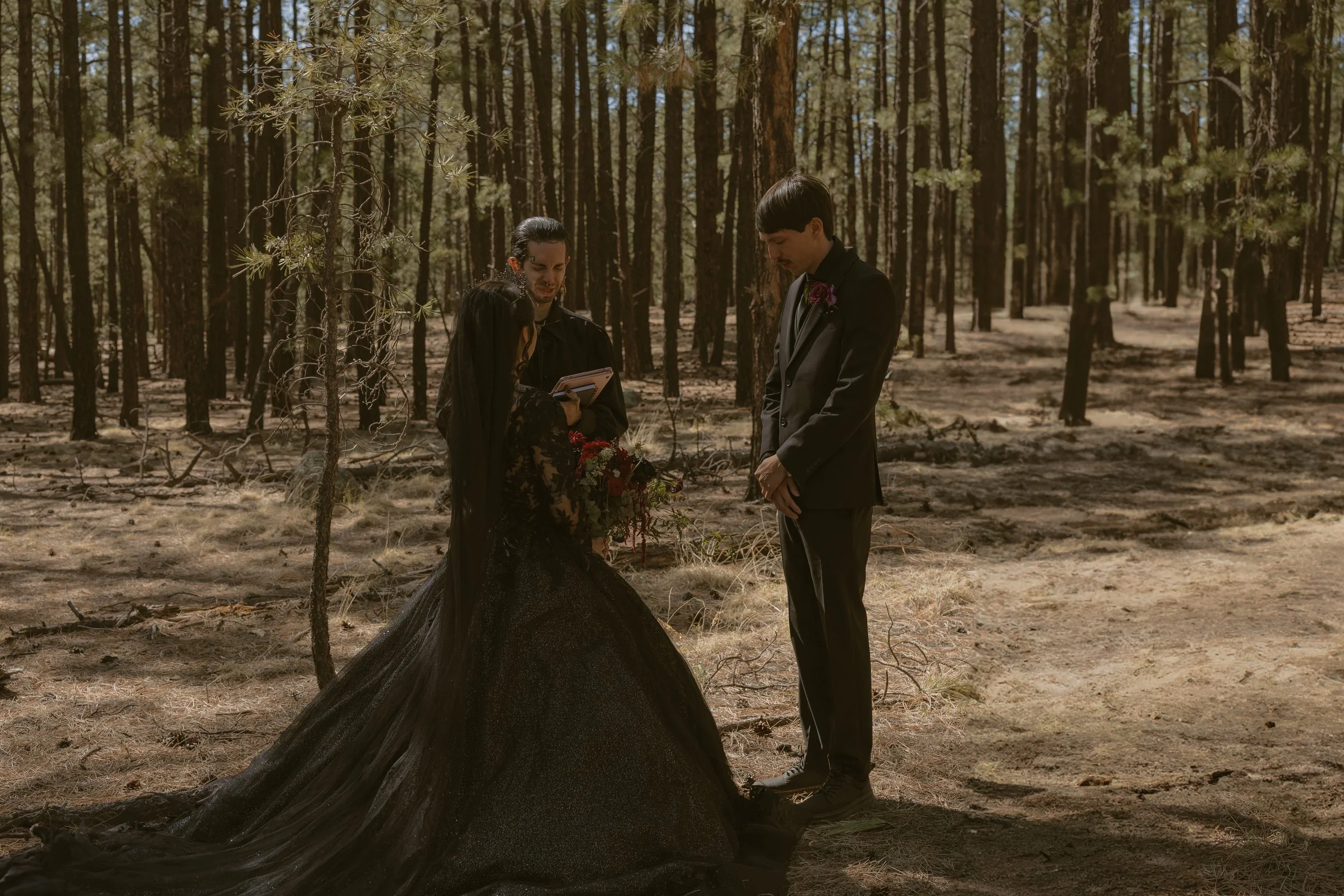 A wedding ceremony in a forest with a bride in a black gown holding a bouquet and two men in suits standing before her, one with a notepad, during daytime.
