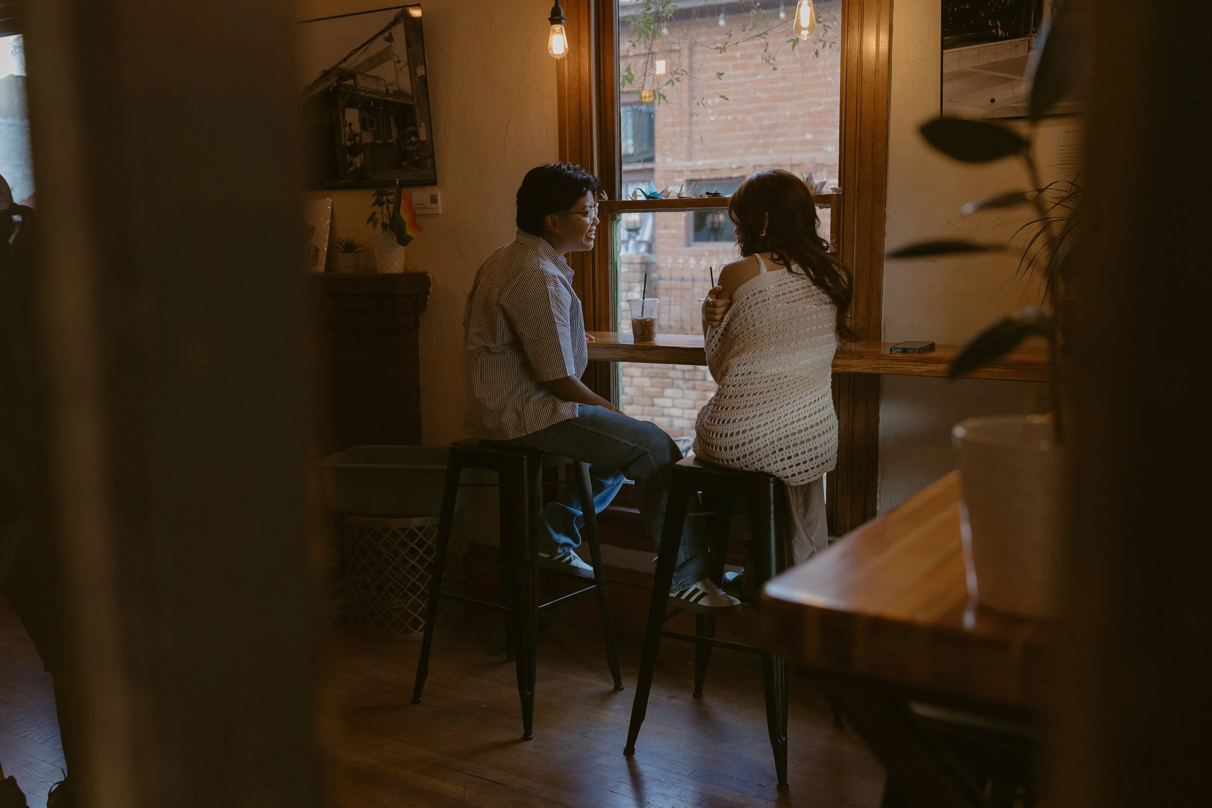 Two people are sitting at a window bench inside a cozy café, having a conversation while enjoying drinks.