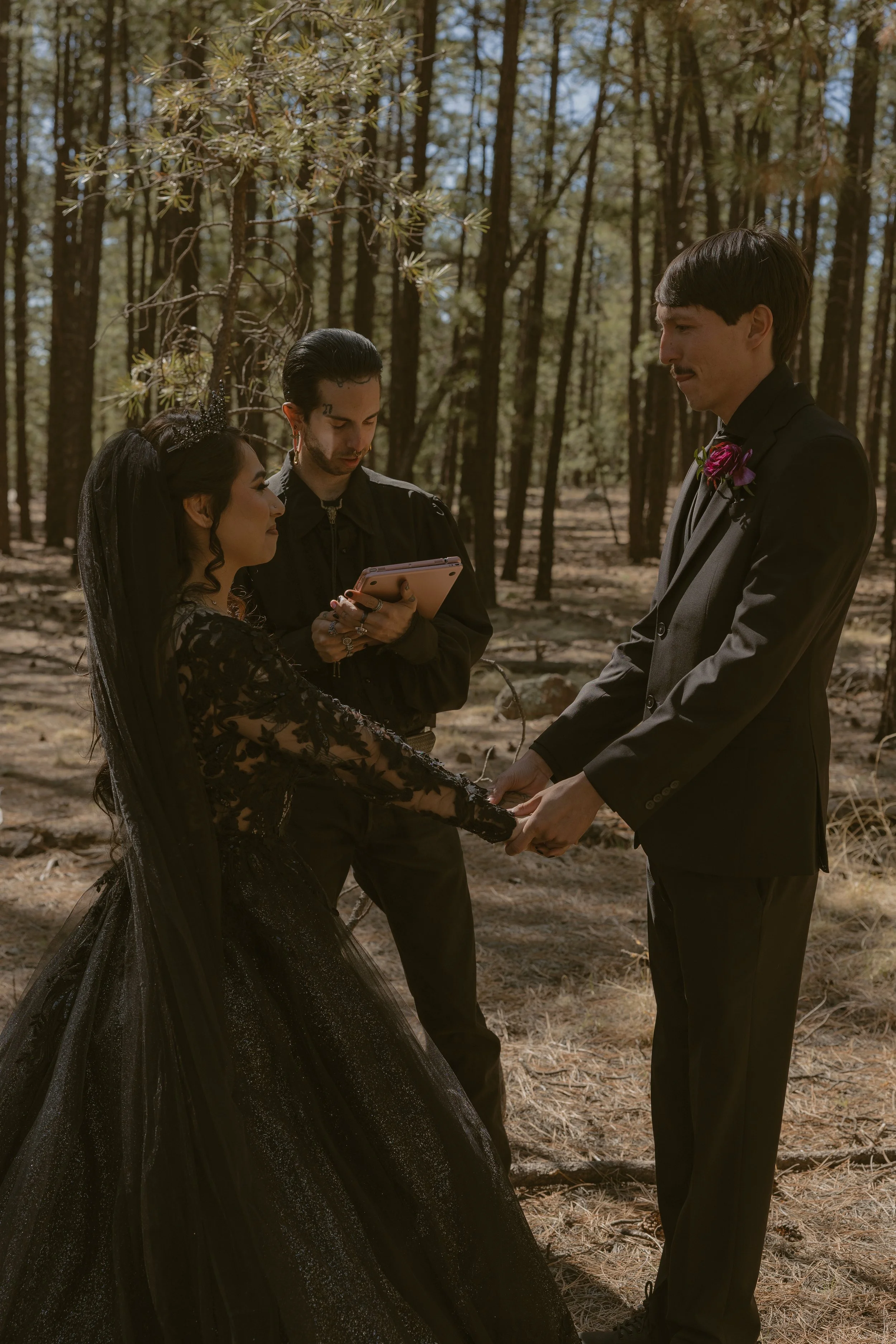 A couple getting married in a forest, holding hands, with a person officiating the ceremony taking notes or reading from a notepad.