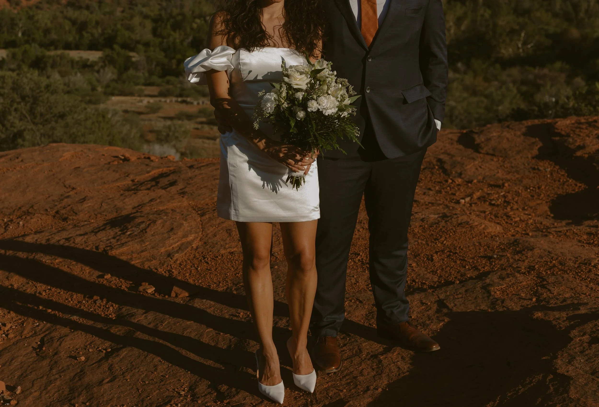 A woman in a short white dress with puff sleeves and white high heels holding a bouquet of white flowers and greenery, standing next to a man in a dark suit and brown shoes, on a rocky outdoor landscape during sunset.