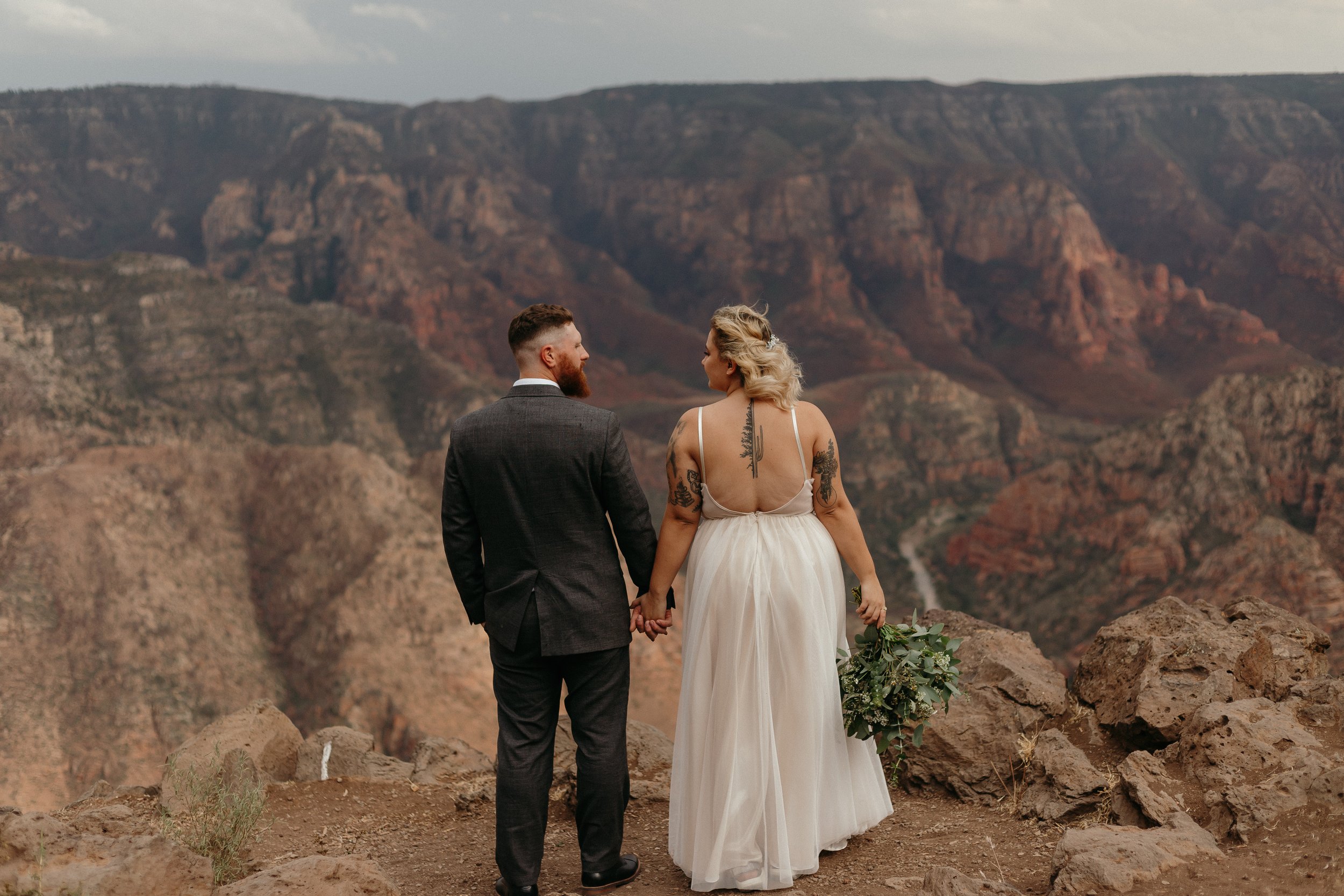 Bride and groom holding hands, standing on rocky cliff edge overlooking a canyon, wedding dress and bouquet.