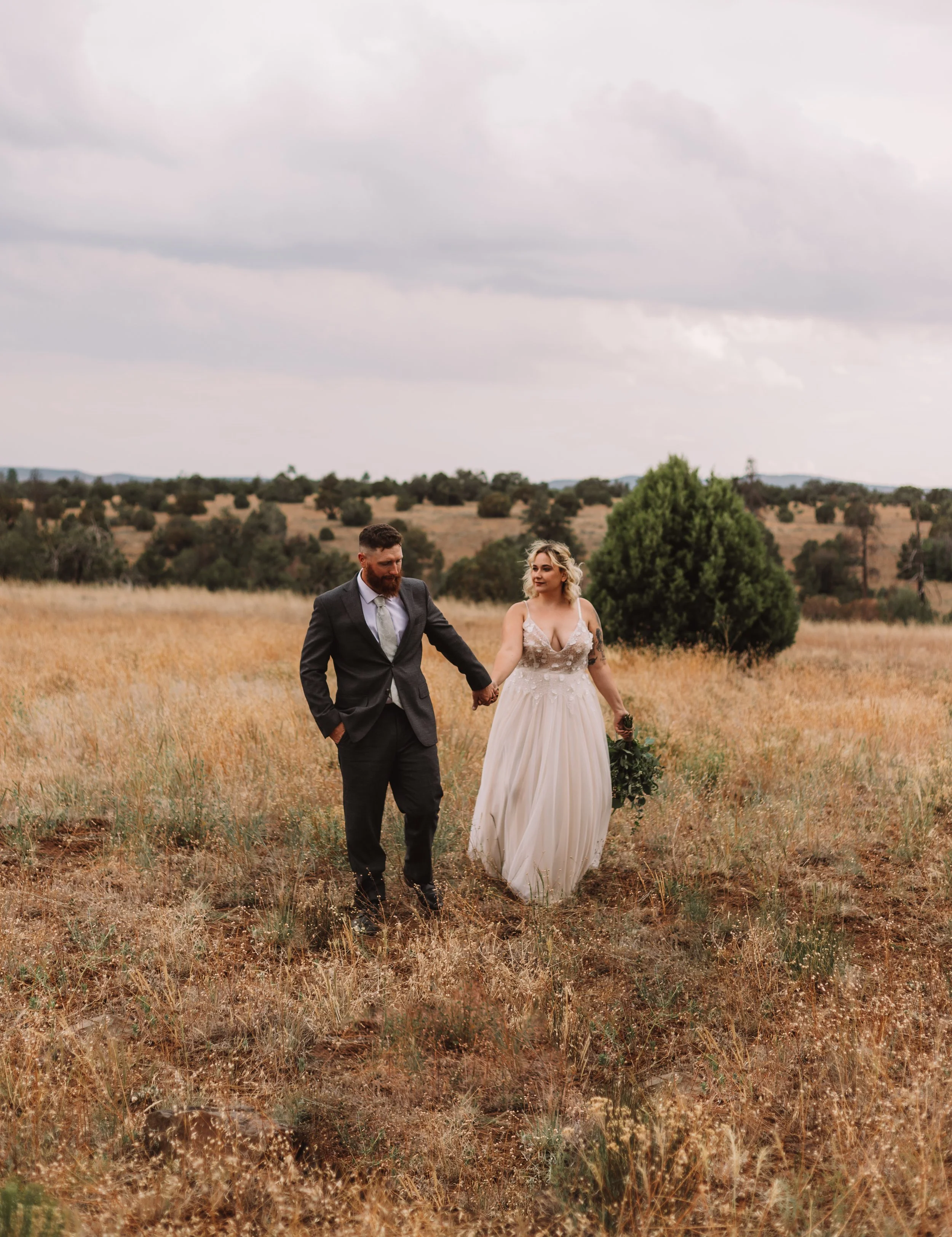 Bride and groom walking in a field of grass under a cloudy sky.