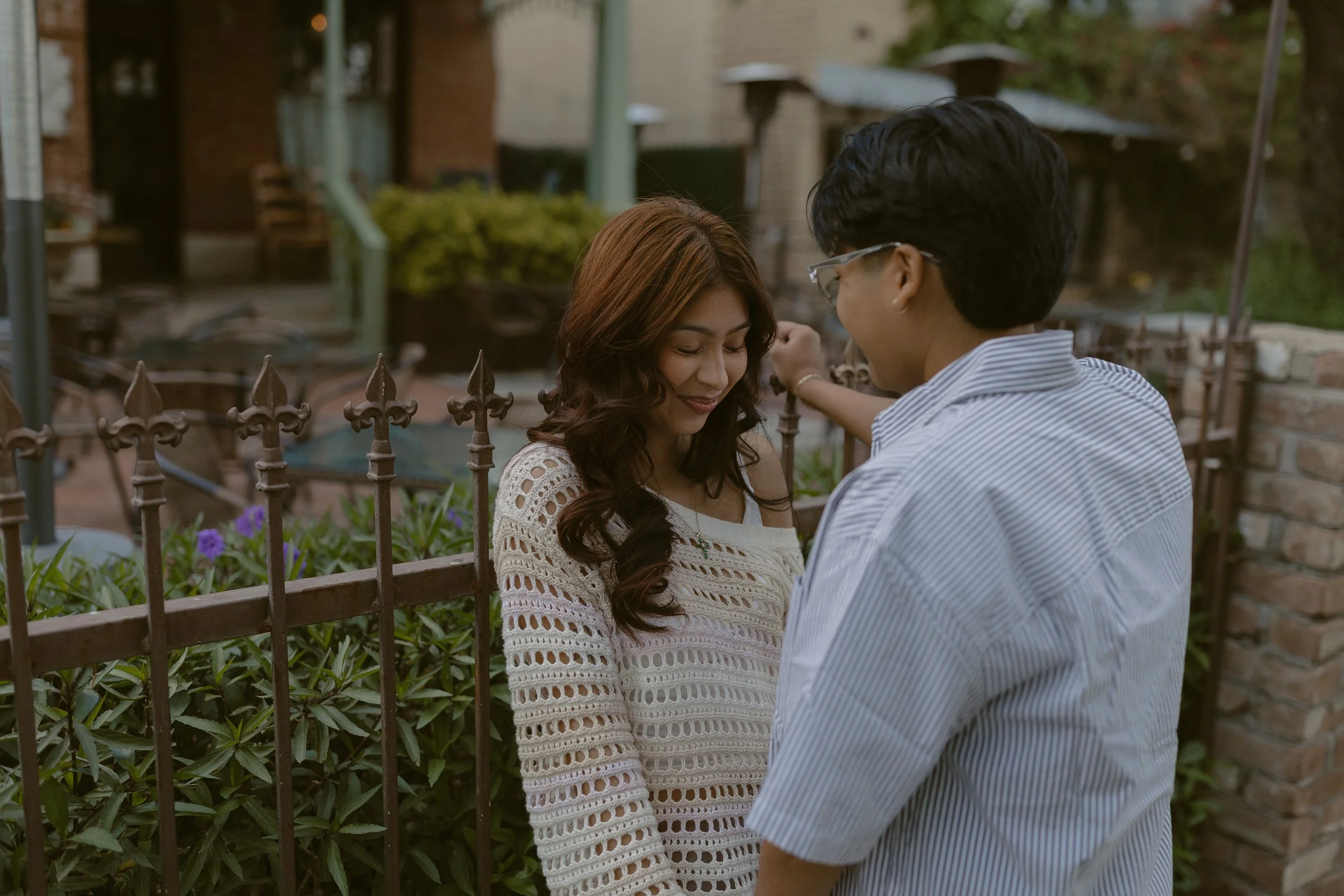 A man and woman stand close together outdoors, with the woman smiling and looking down while the man touches her shoulder, behind a decorative iron fence and greenery.