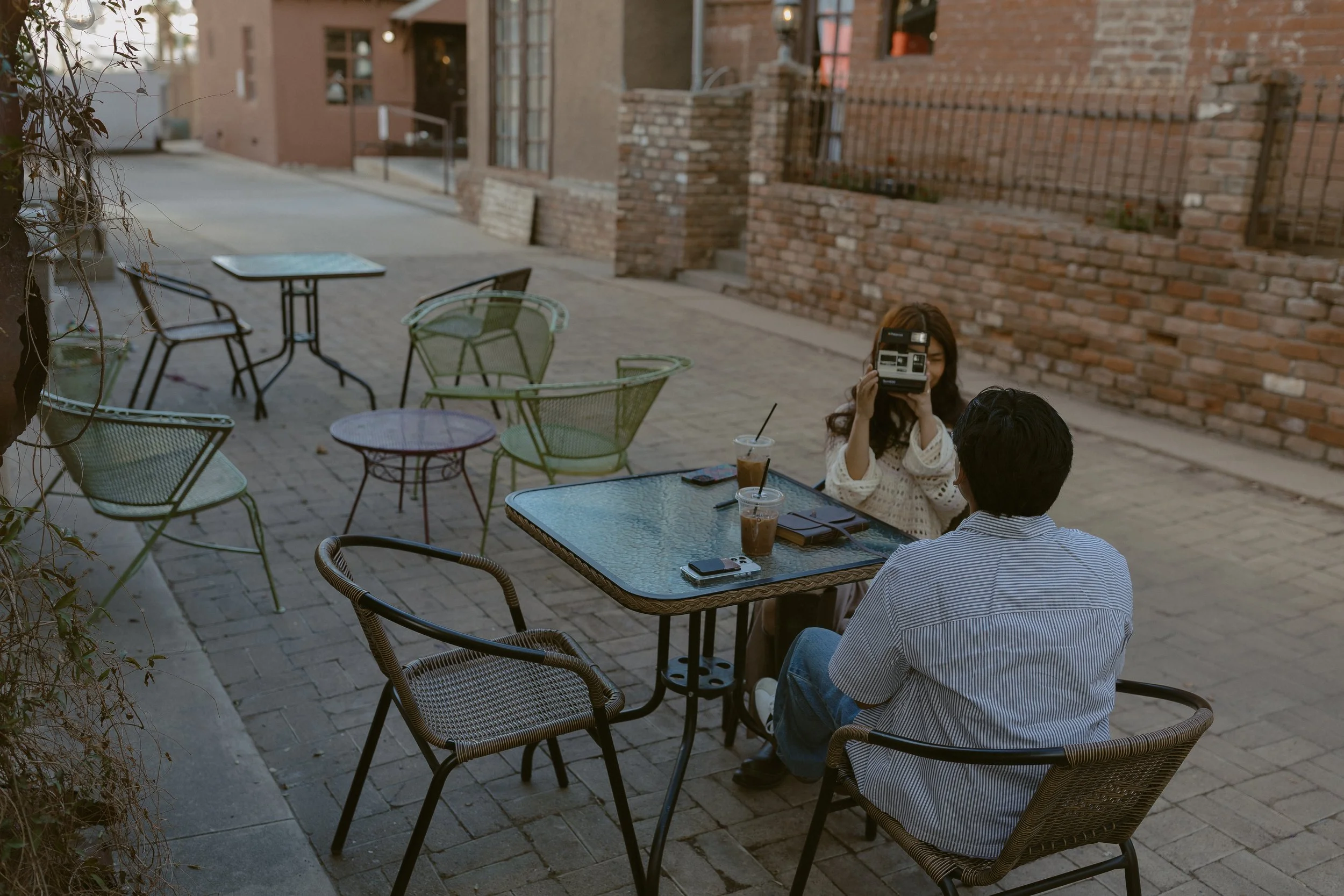 A woman is taking a photo of a man sitting at an outdoor cafe table with sunglasses and drinks on the table, in an alleyway with brick walls and empty chairs and tables.